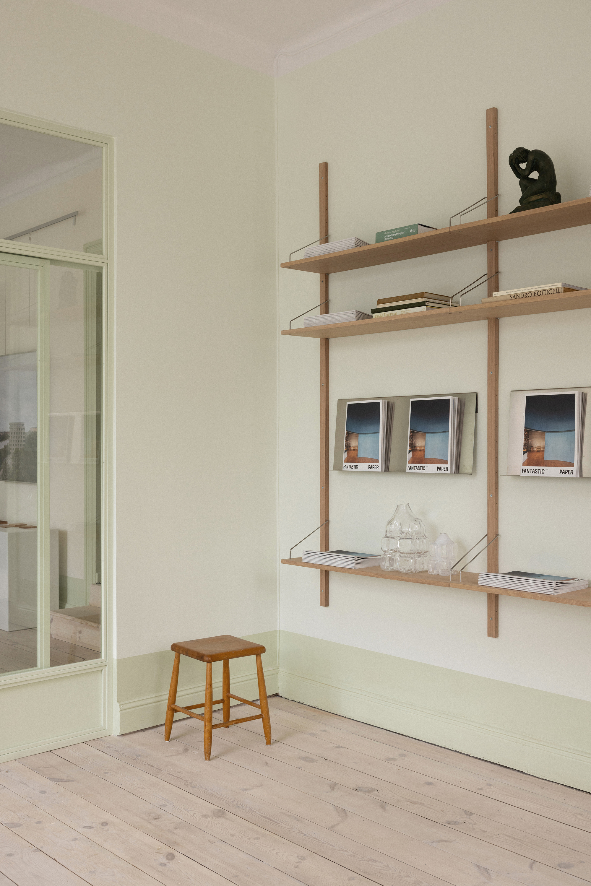 Wall-mounted wooden shelving system with vertical supports displaying books and framed prints beside a low stool