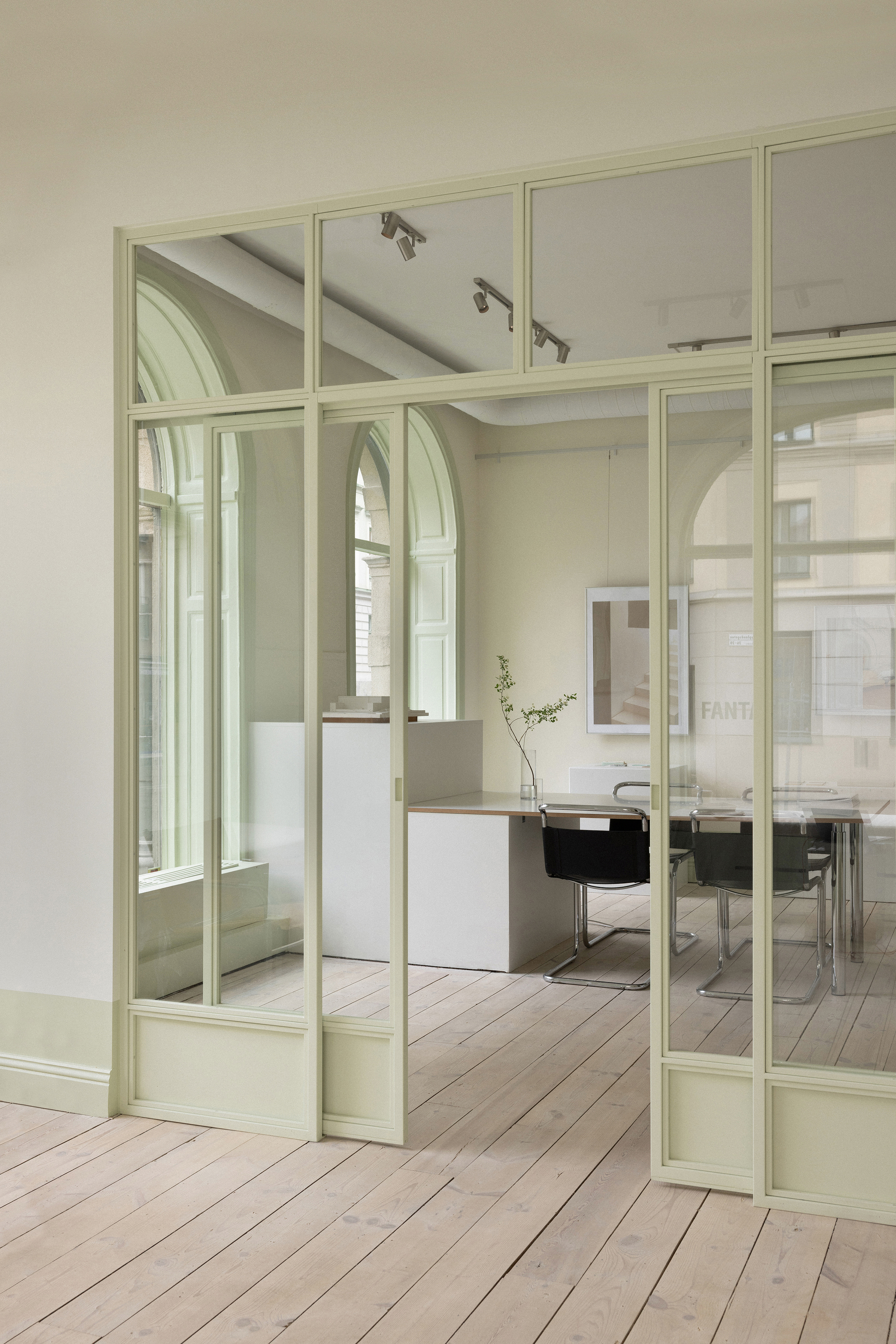 View through the pale green steel and glass partition showing arched windows and track lighting beyond