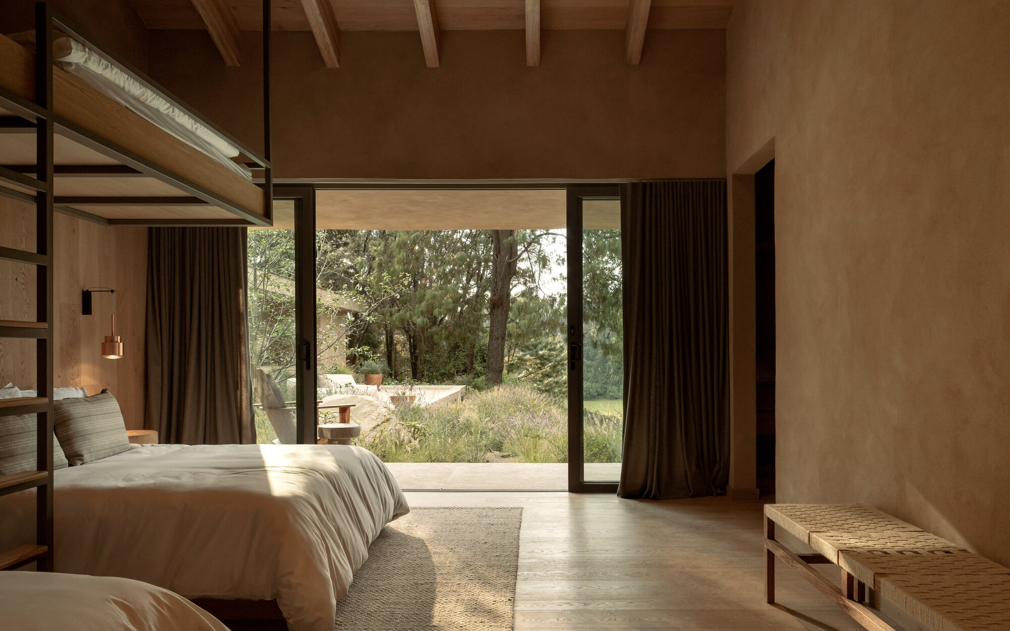 Bedroom with full-height glazing opening to a meadow under dappled morning sunlight