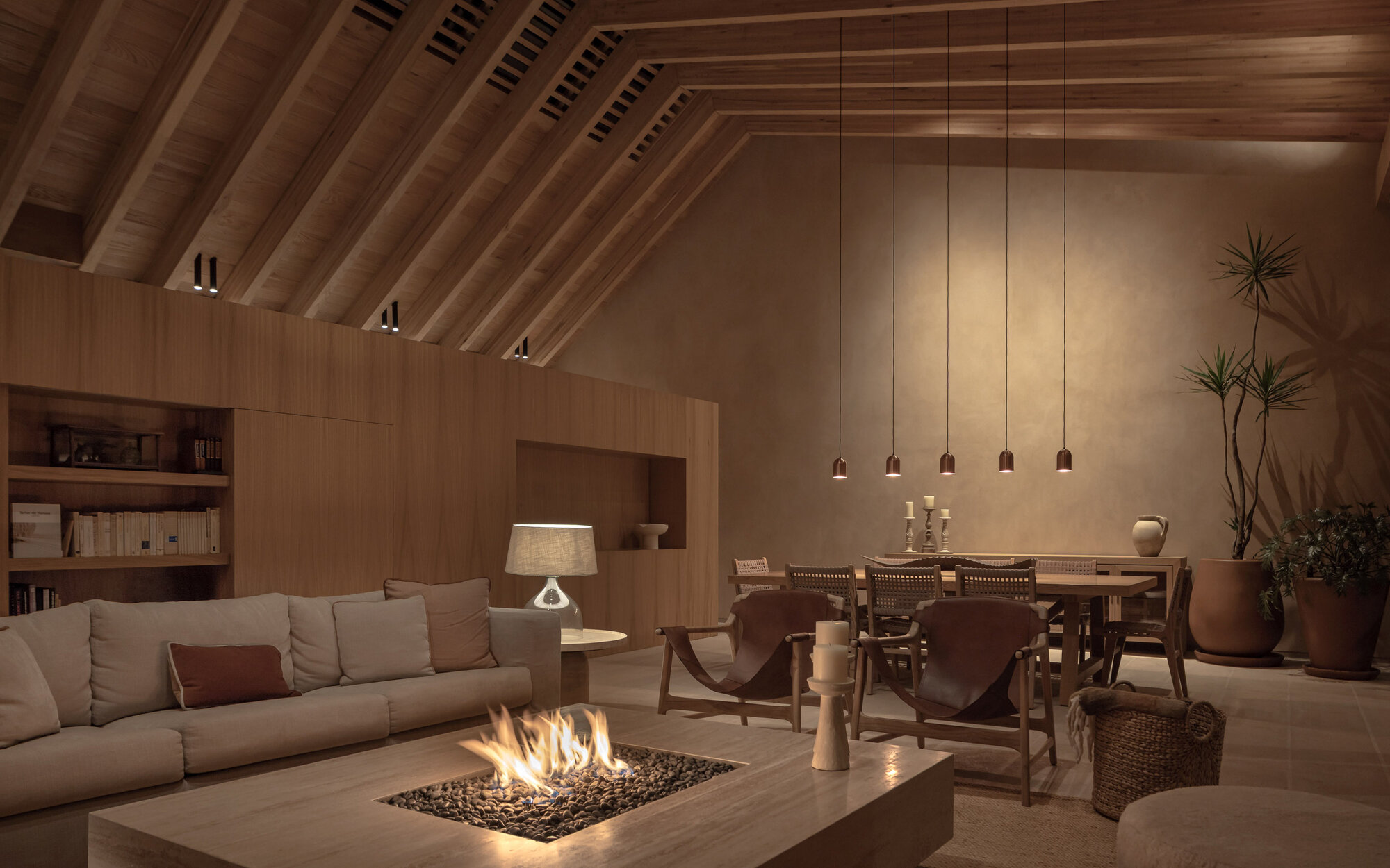 Open-plan living and dining space with exposed timber rafters and a central fire pit at dusk