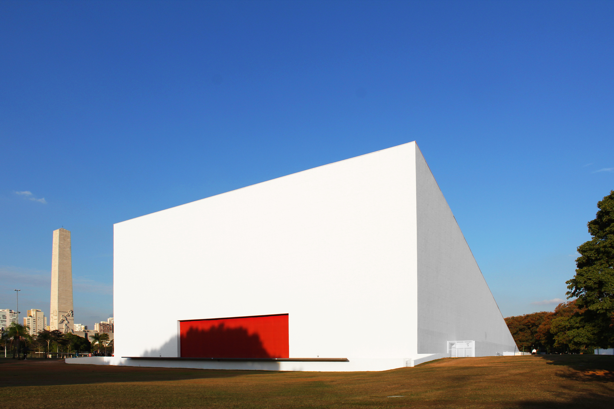 White triangular volume with a recessed red-lit entrance opening on a grass field under clear blue sky