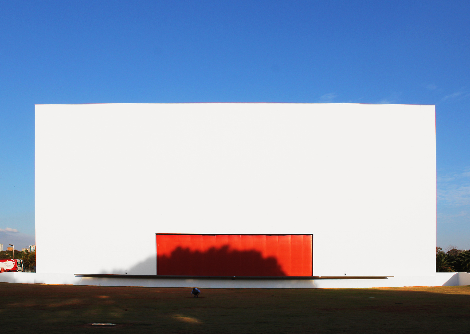 White cubic facade with a red backlit rectangular opening casting a shadow on the lawn at dusk