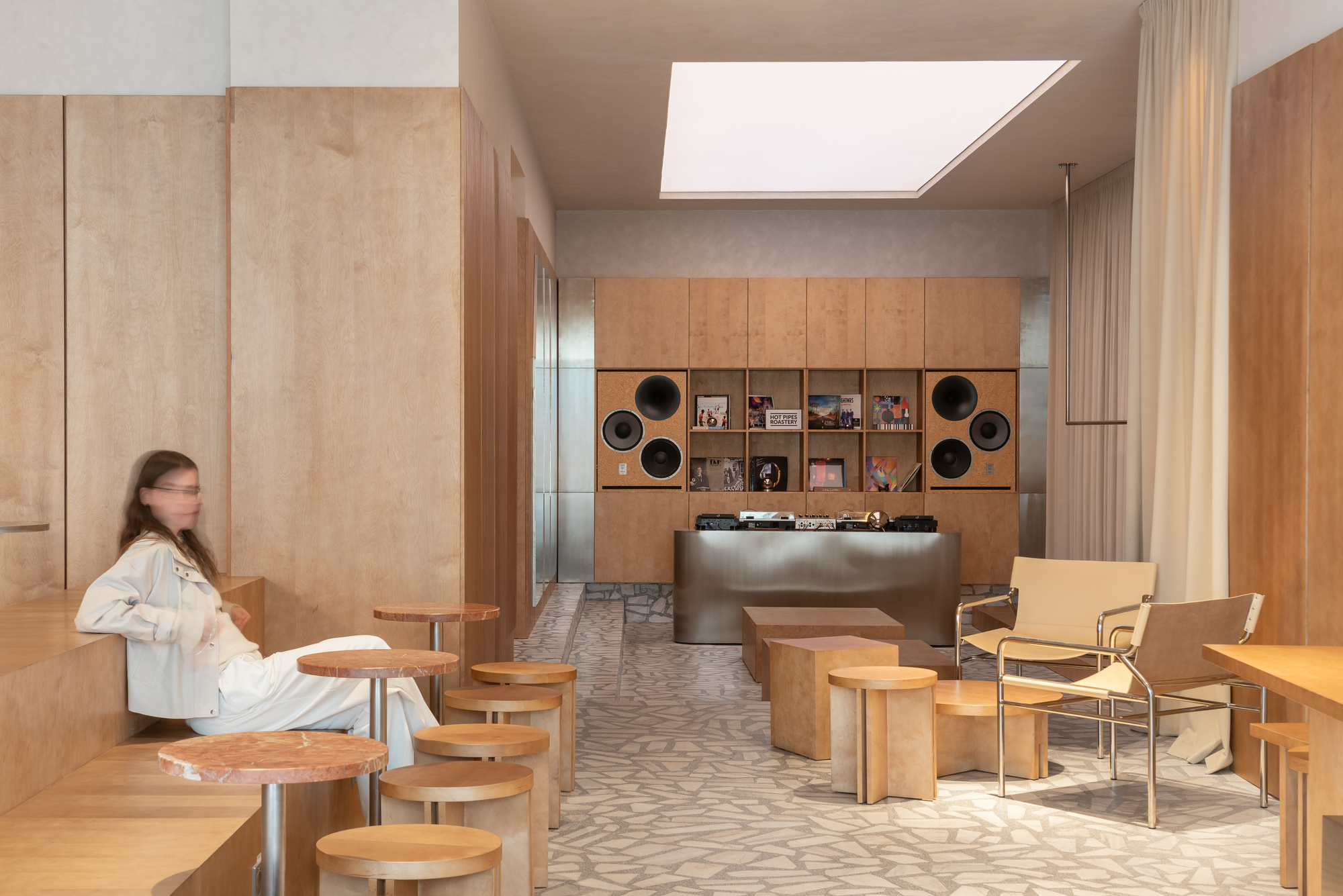 Interior cafe with plywood cabinetry and embedded speakers illuminated by a large skylight overhead
