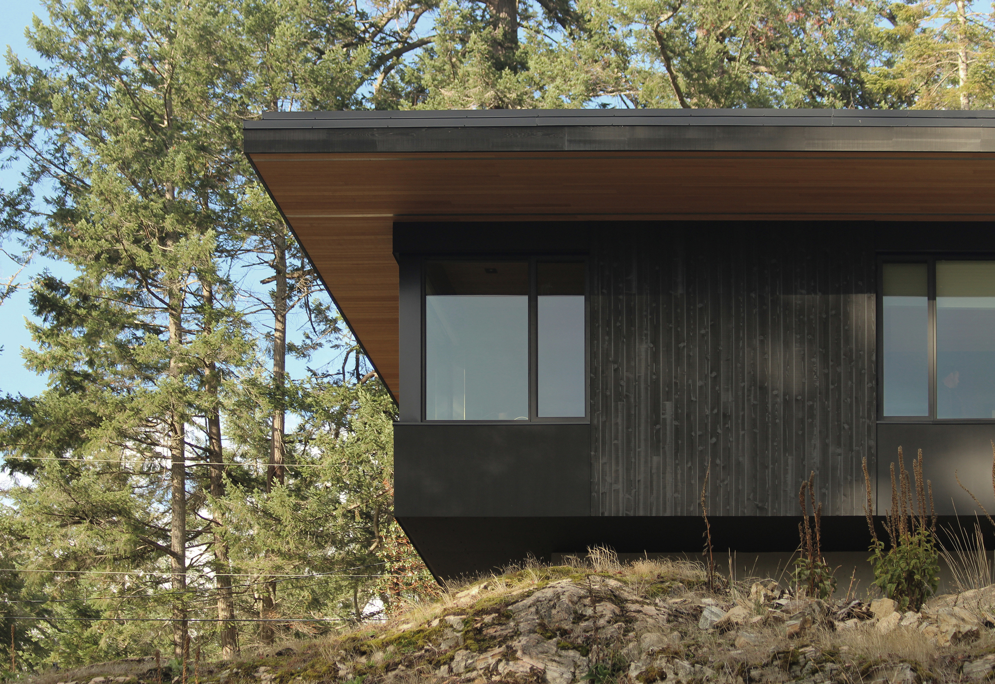 Cantilevered roof with cedar soffit extending over charred wood cladding and hillside boulders