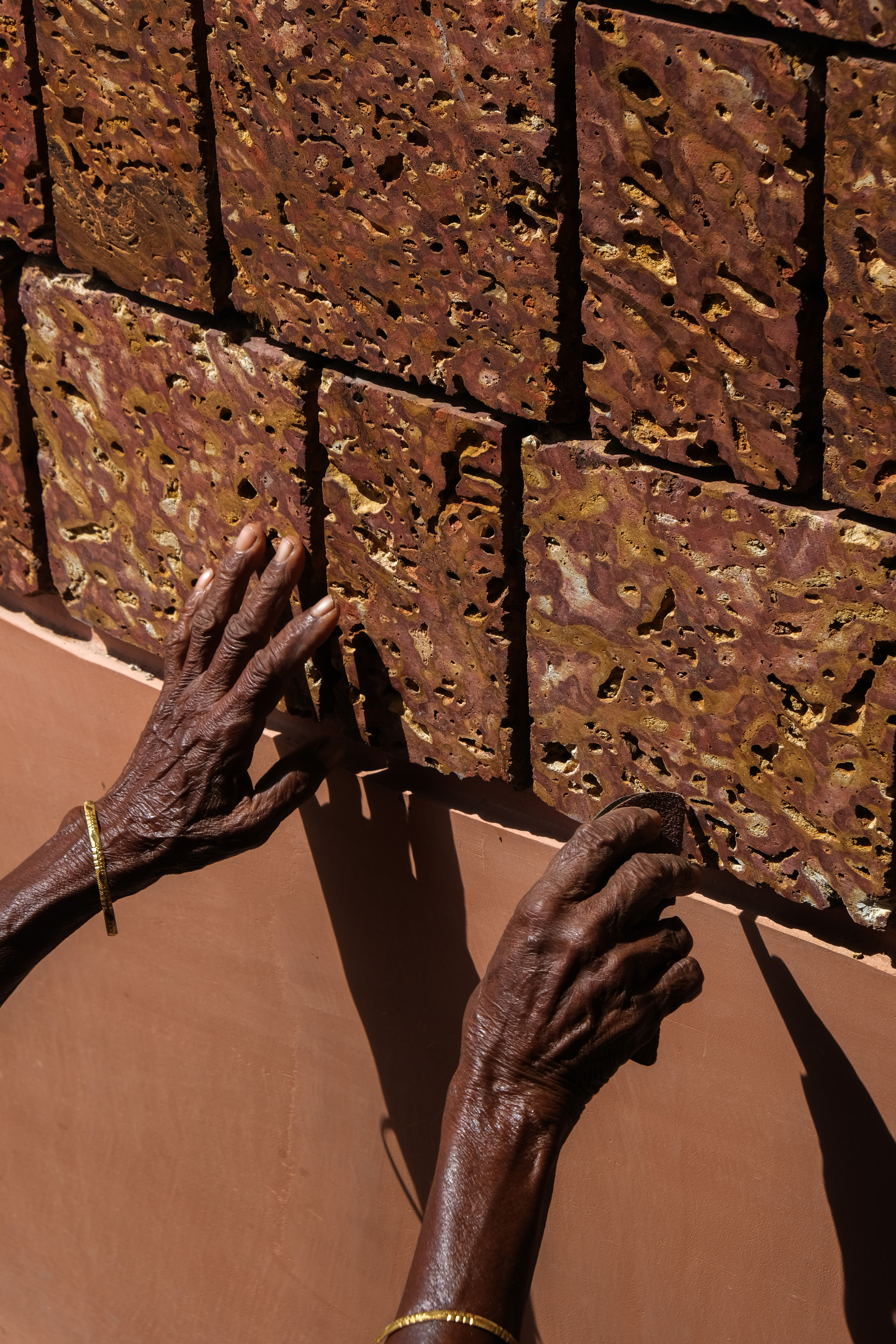 Close-up of hands touching textured laterite brick wall showing irregular surface and deep mortar joints in sunlight