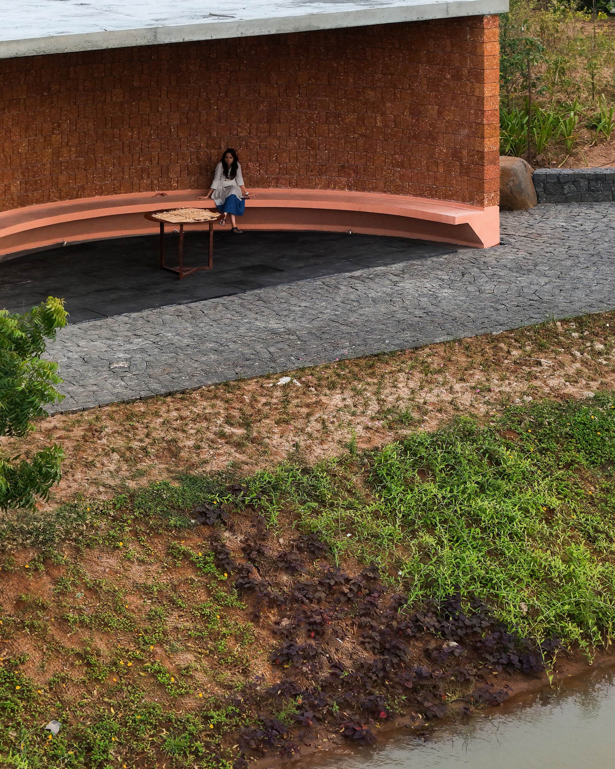 Curved terracotta bench against laterite brick wall with seated figure on dark stone paving beside pond