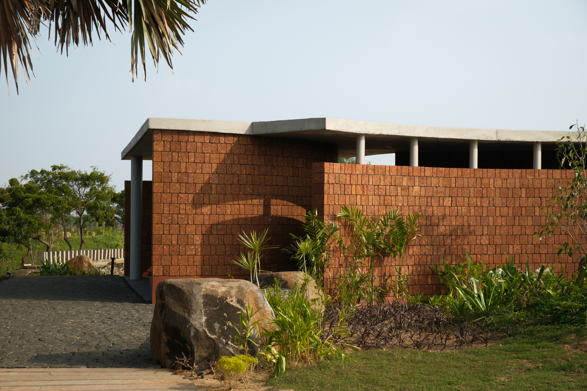 Red laterite brick wall with arched entry opening set among palm fronds and planted boulders