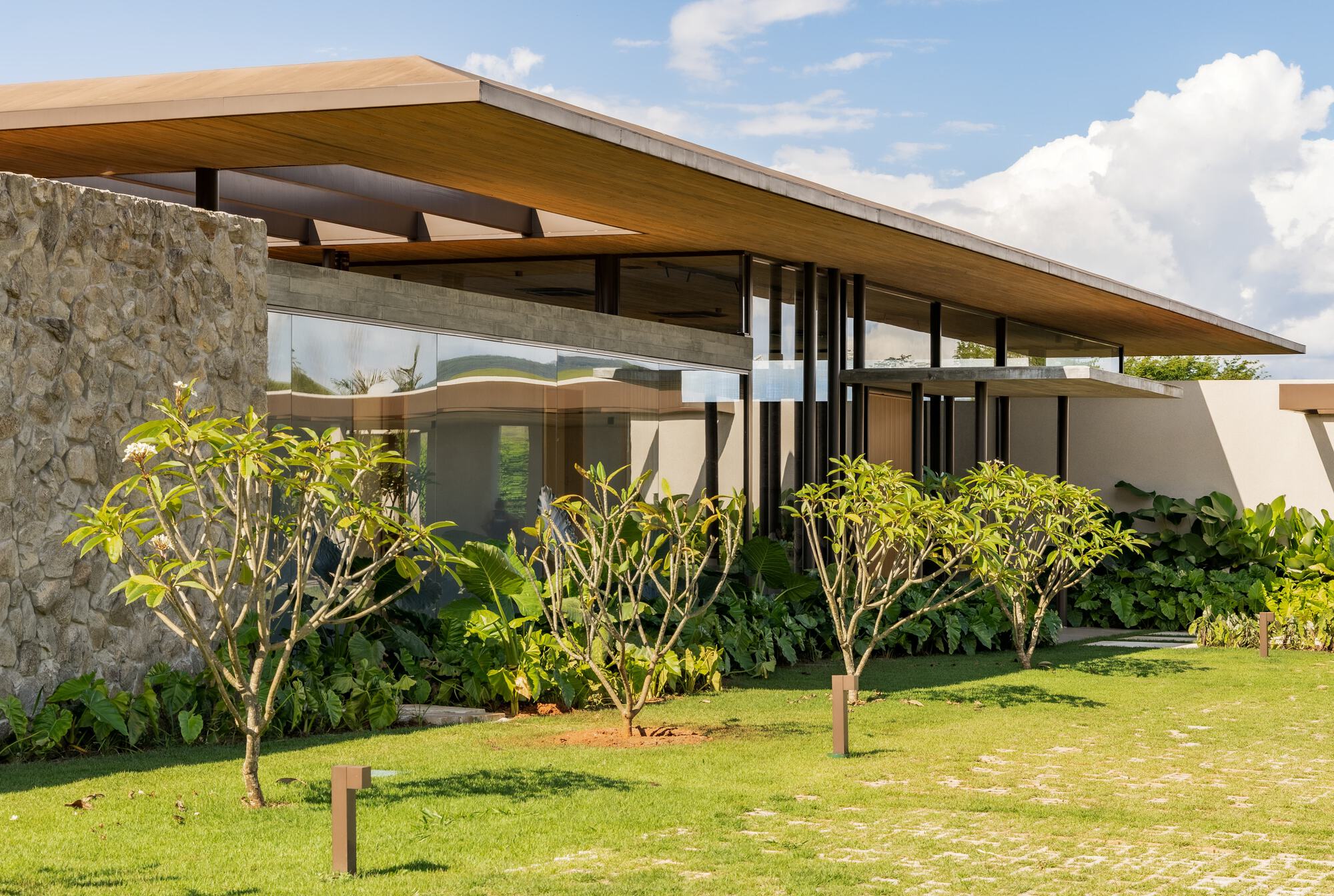 Cantilevered timber roof plane over stone and glass volumes edged by lawn and planted beds