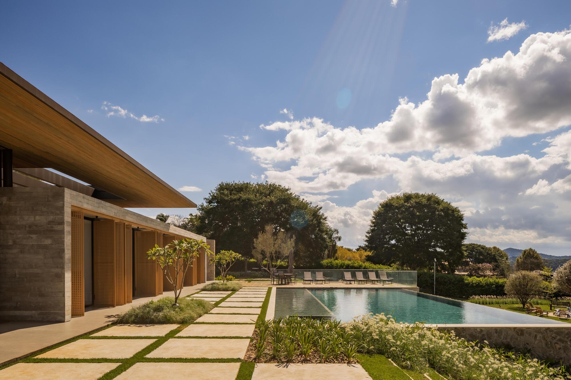 Pool deck with stone pavers set in lawn leading to concrete volume with timber soffit under cloudscape
