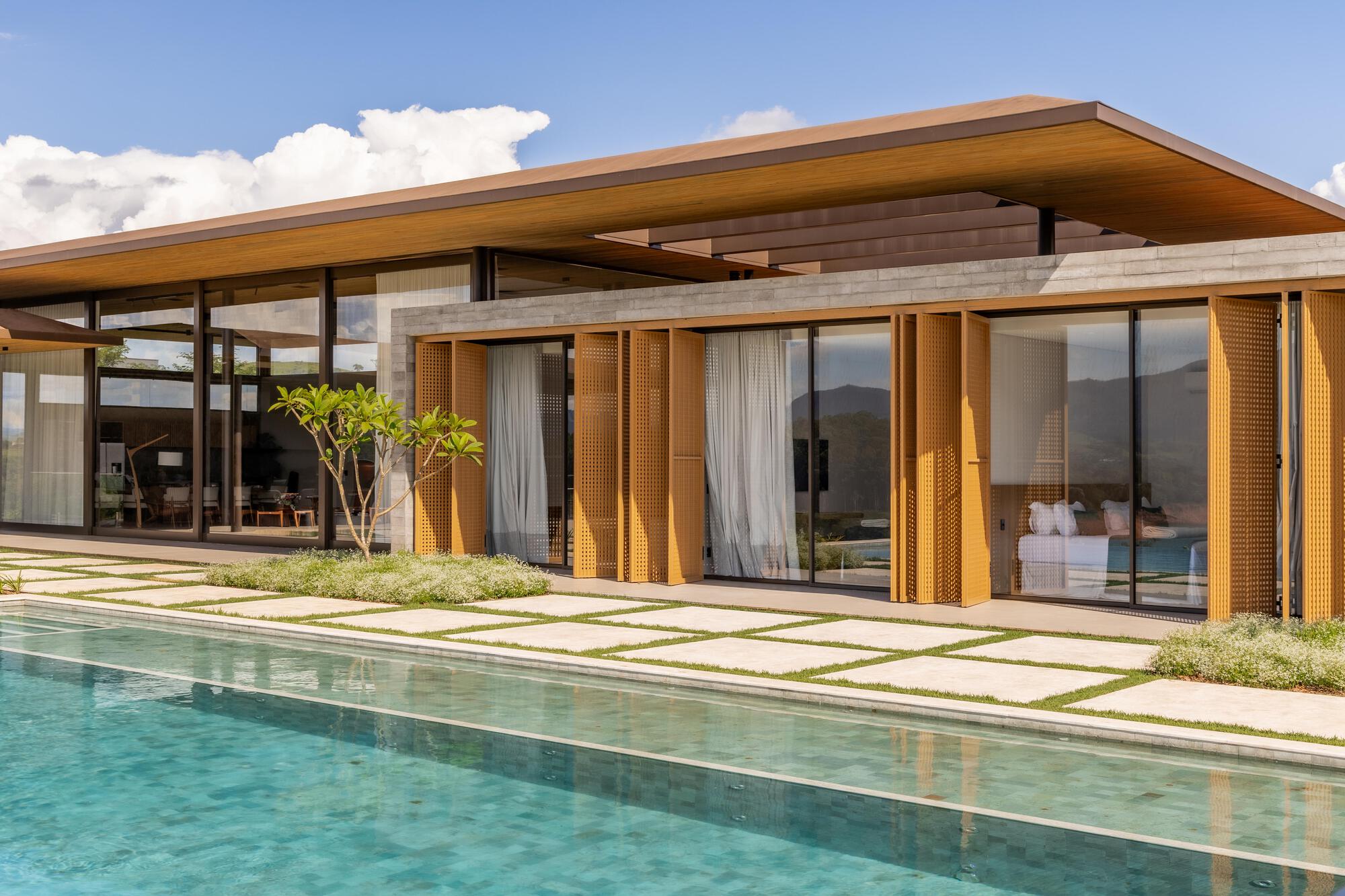 Cantilevered timber roof over glass-walled pavilion with perforated wooden screens beside the swimming pool
