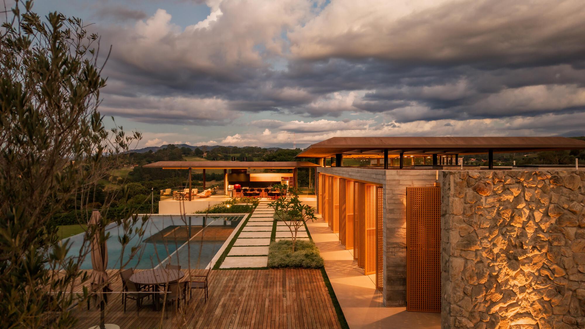 Timber deck bridge connecting the pool pavilion and stone-clad volume under stormy skies