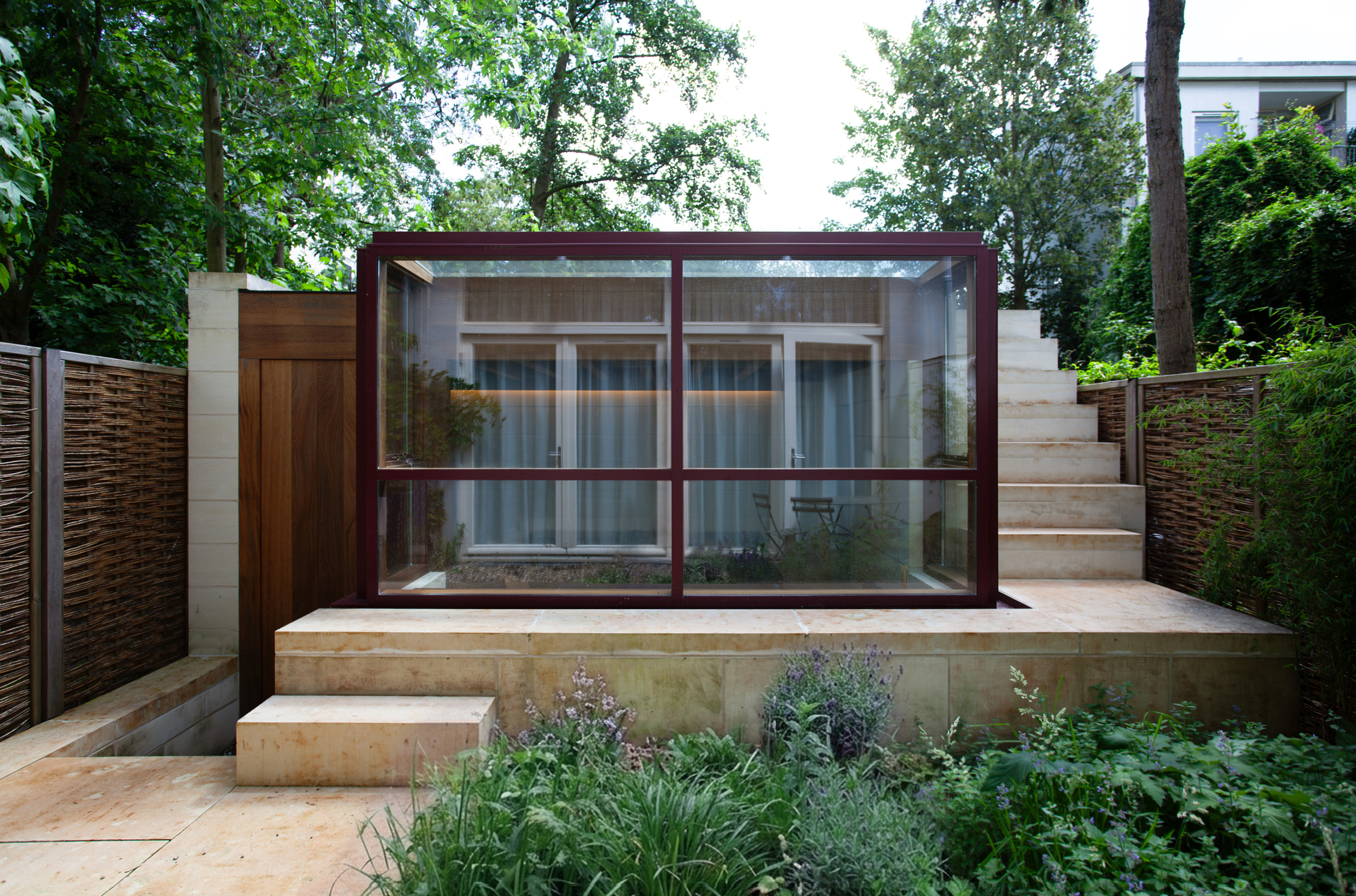Garden view of the glass-walled pavilion with red steel frame sitting on a stone plinth surrounded by planting
