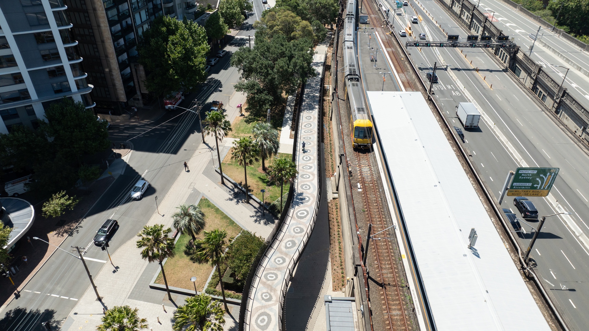 Overhead view of the elevated railway platform with white canopy roof running between landscaped pathways and highway