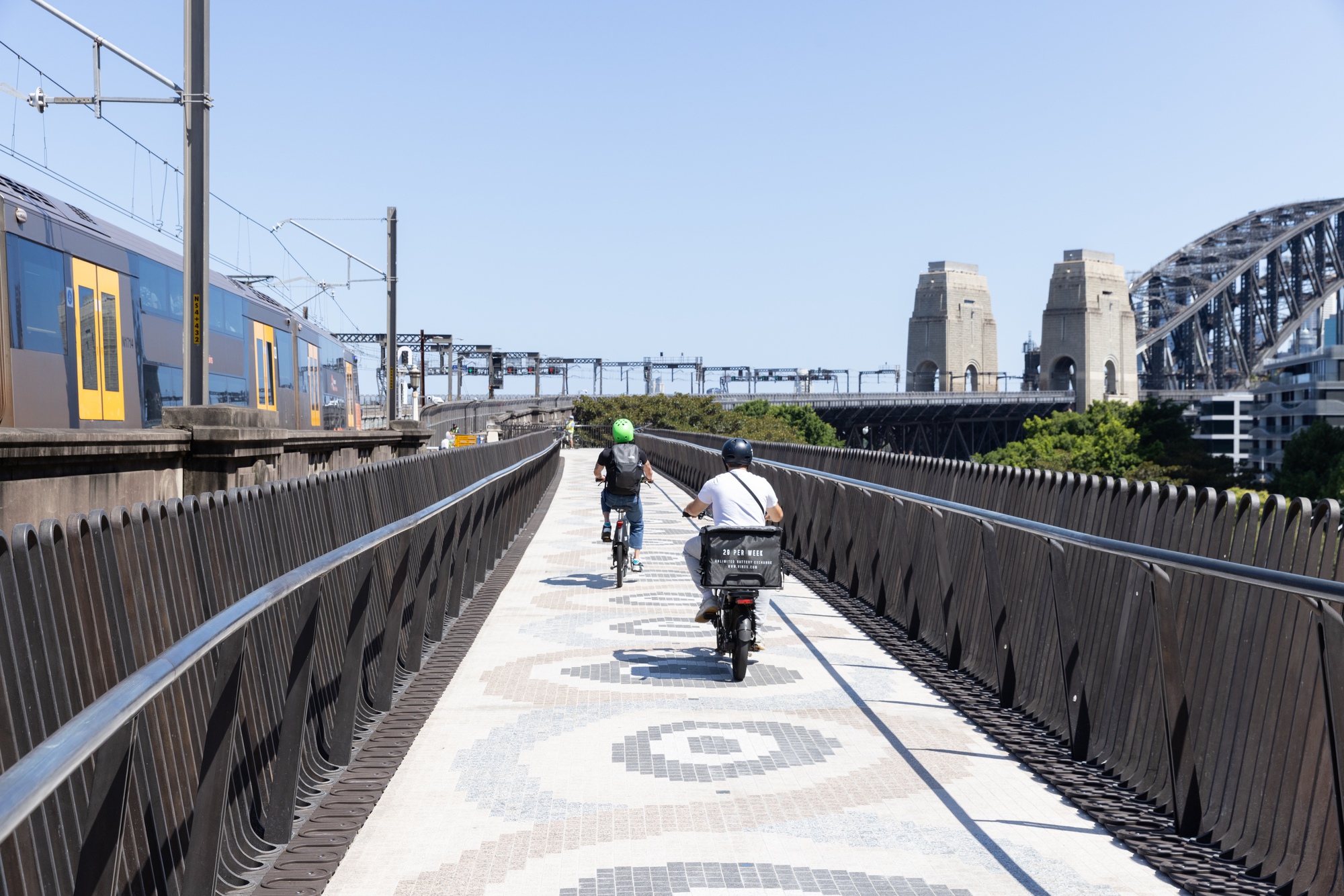 Pedestrian and cycle bridge with patterned concrete paving and vertical metal railings under clear afternoon sky
