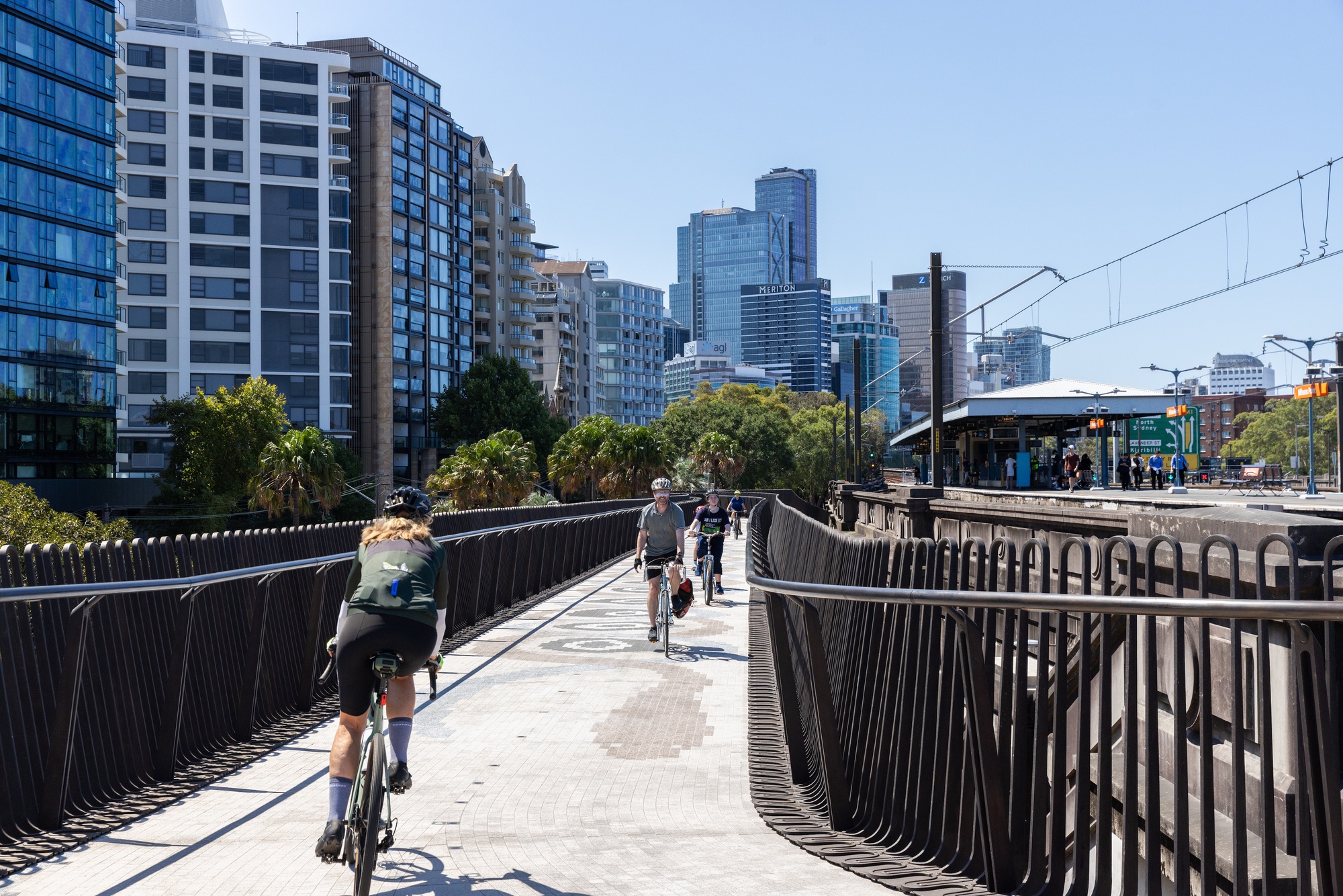 Cyclists and pedestrians on the undulating bridge deck with vertical metal balusters and urban skyline beyond