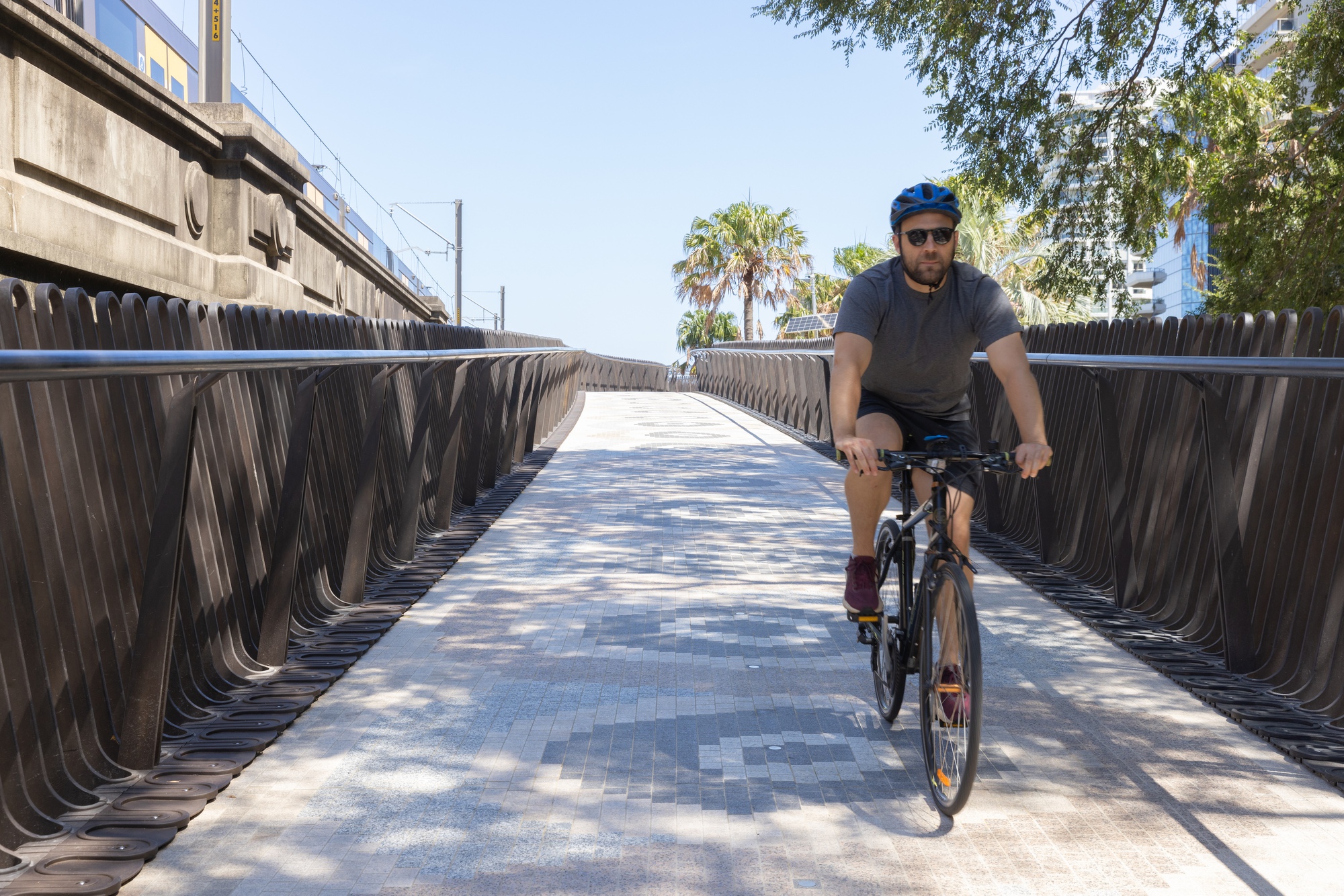 Pedestrian bridge with curved metal railings and patterned paving as a cyclist approaches under palms