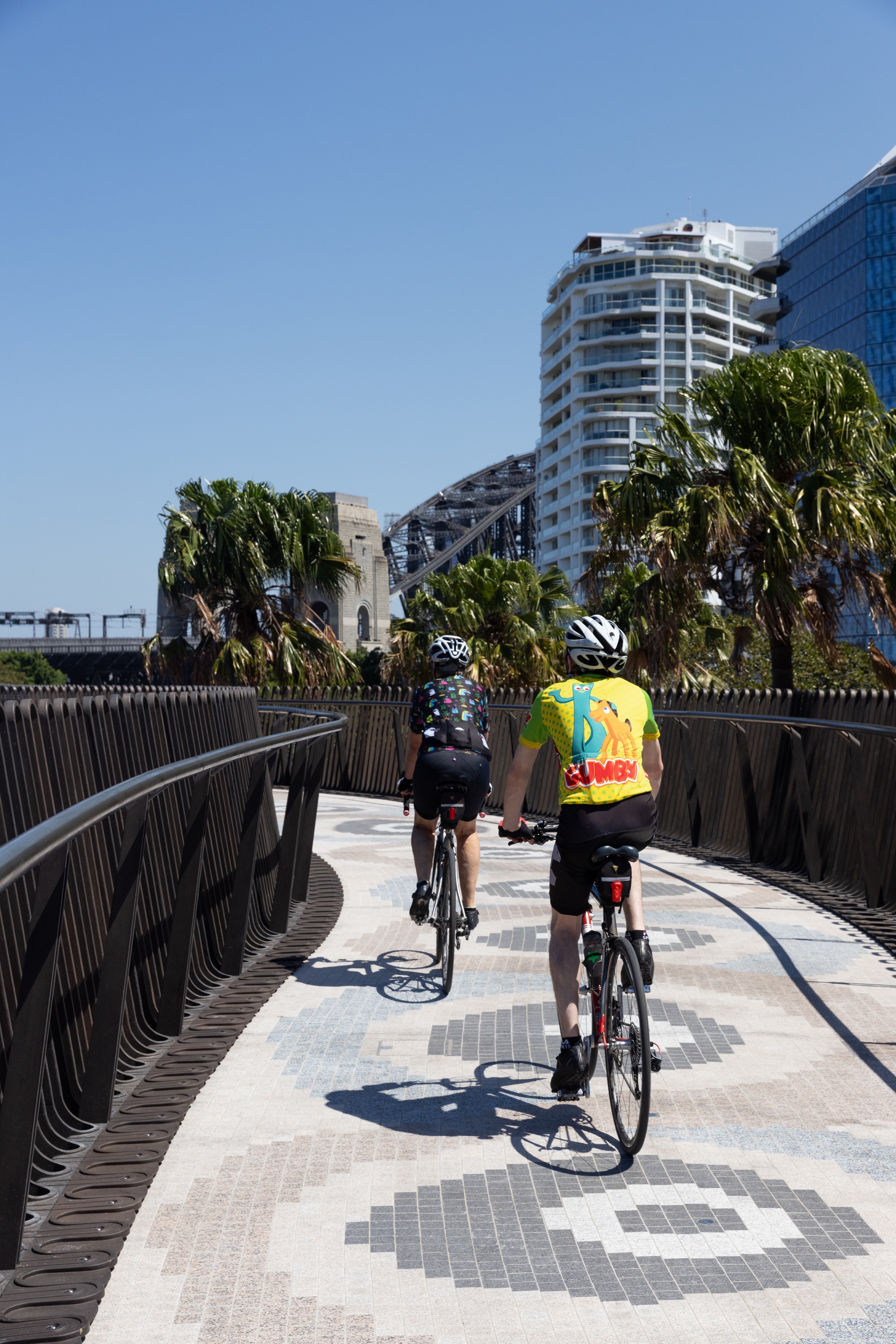 Elevated pathway with dark vertical balustrade and cyclists riding toward residential towers in sunlight
