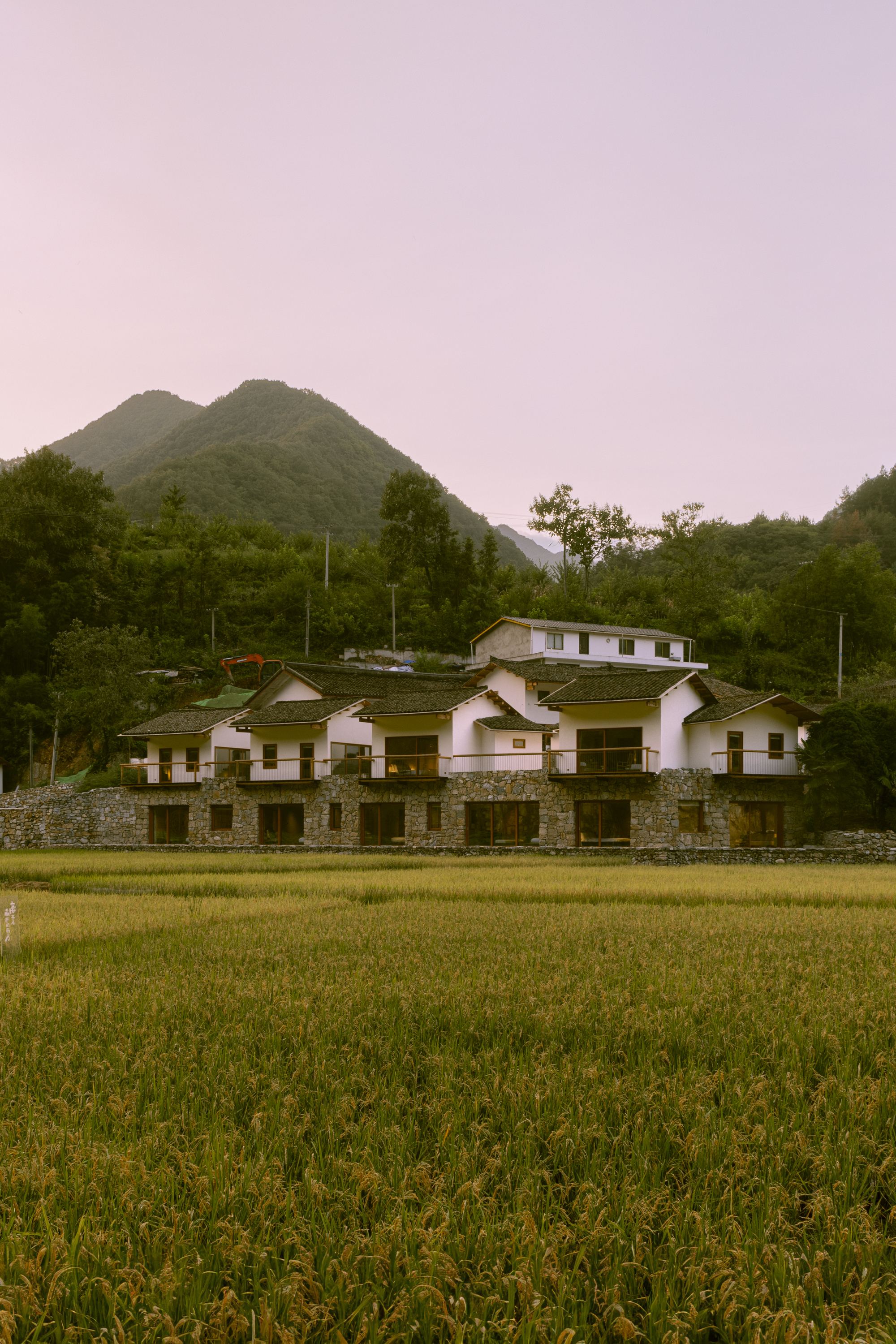 Rear elevation with tile roofs and stone plinth wall set against golden rice fields and forested peaks