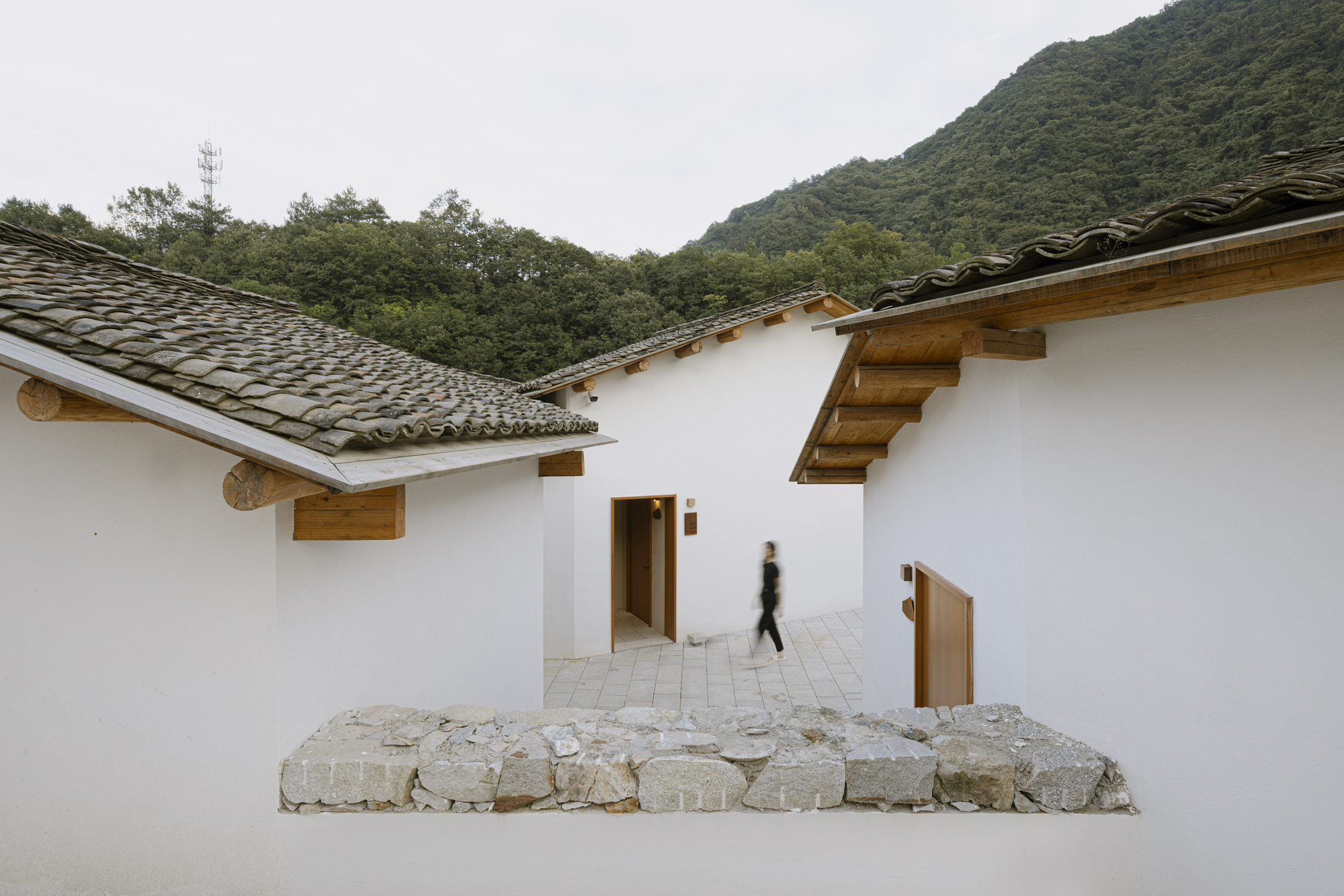 Courtyard passage with white plastered walls, timber eaves, and fieldstone outcrop with a figure passing through