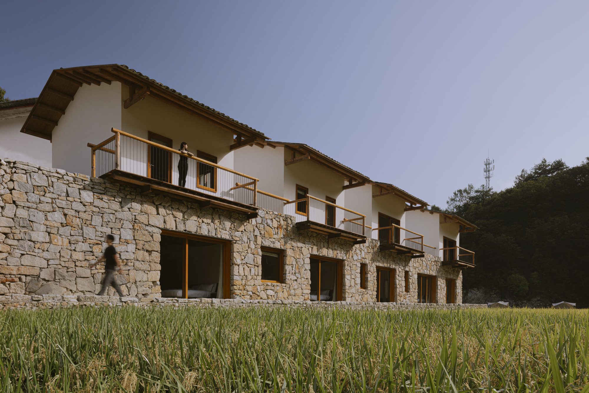 Repeating balcony modules with timber railings along a stone masonry base as figures move through the scene