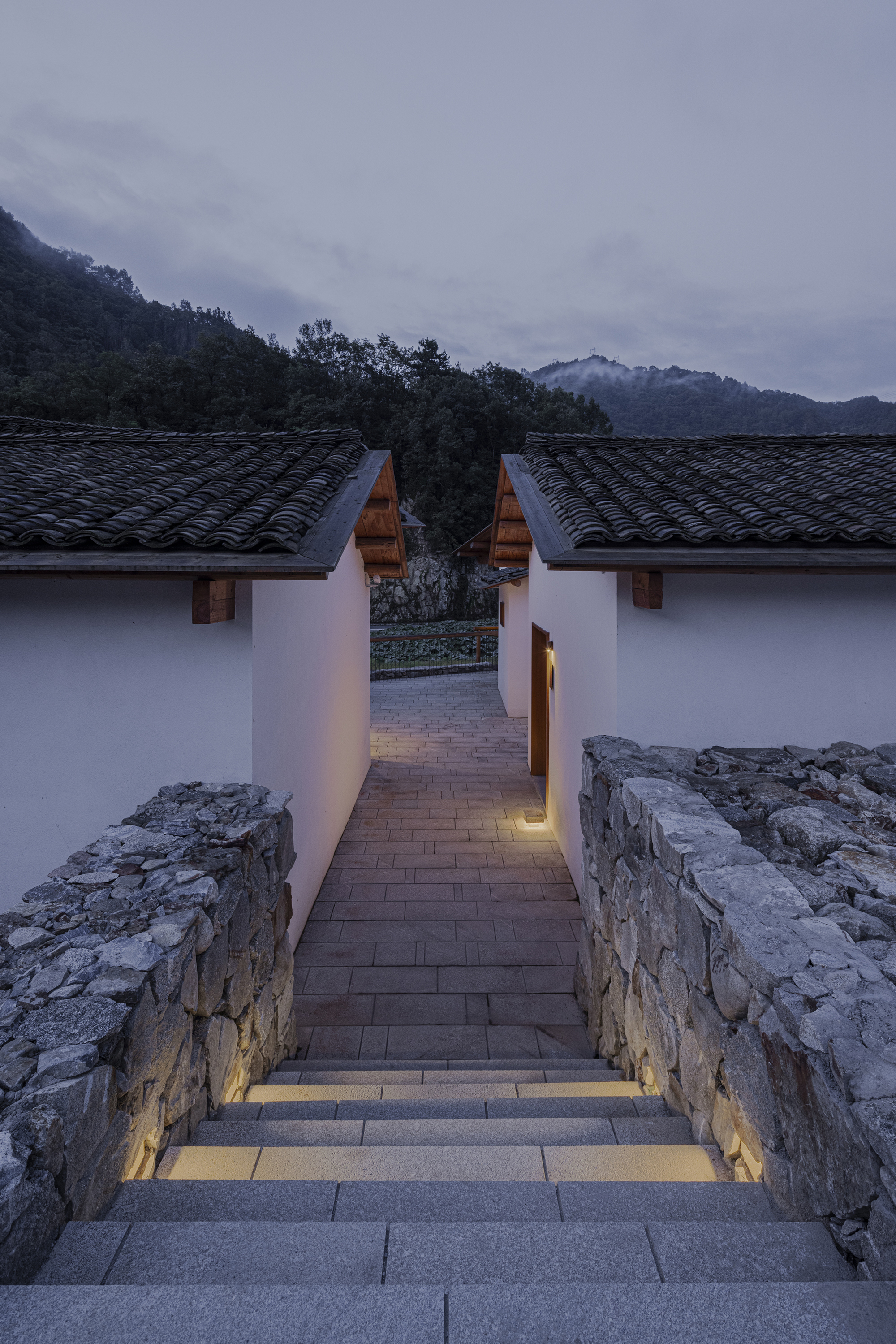 Stone staircase with recessed lighting descending between white walls toward tiled roofs at dusk