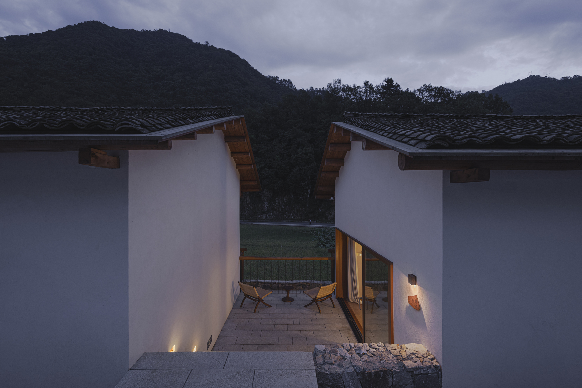 Entry courtyard between two white volumes with timber eaves and uplighting against mountain backdrop at twilight