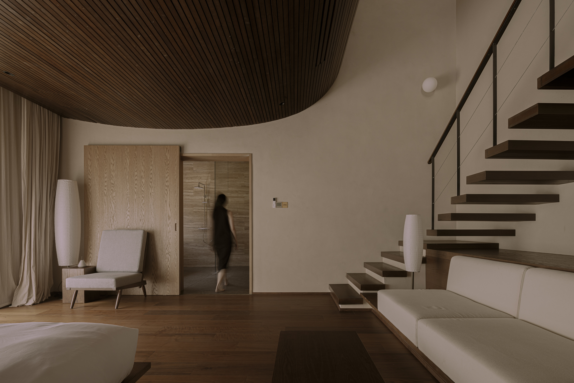Interior space with cantilevered timber stair and curved slatted ceiling as figure walks through doorway