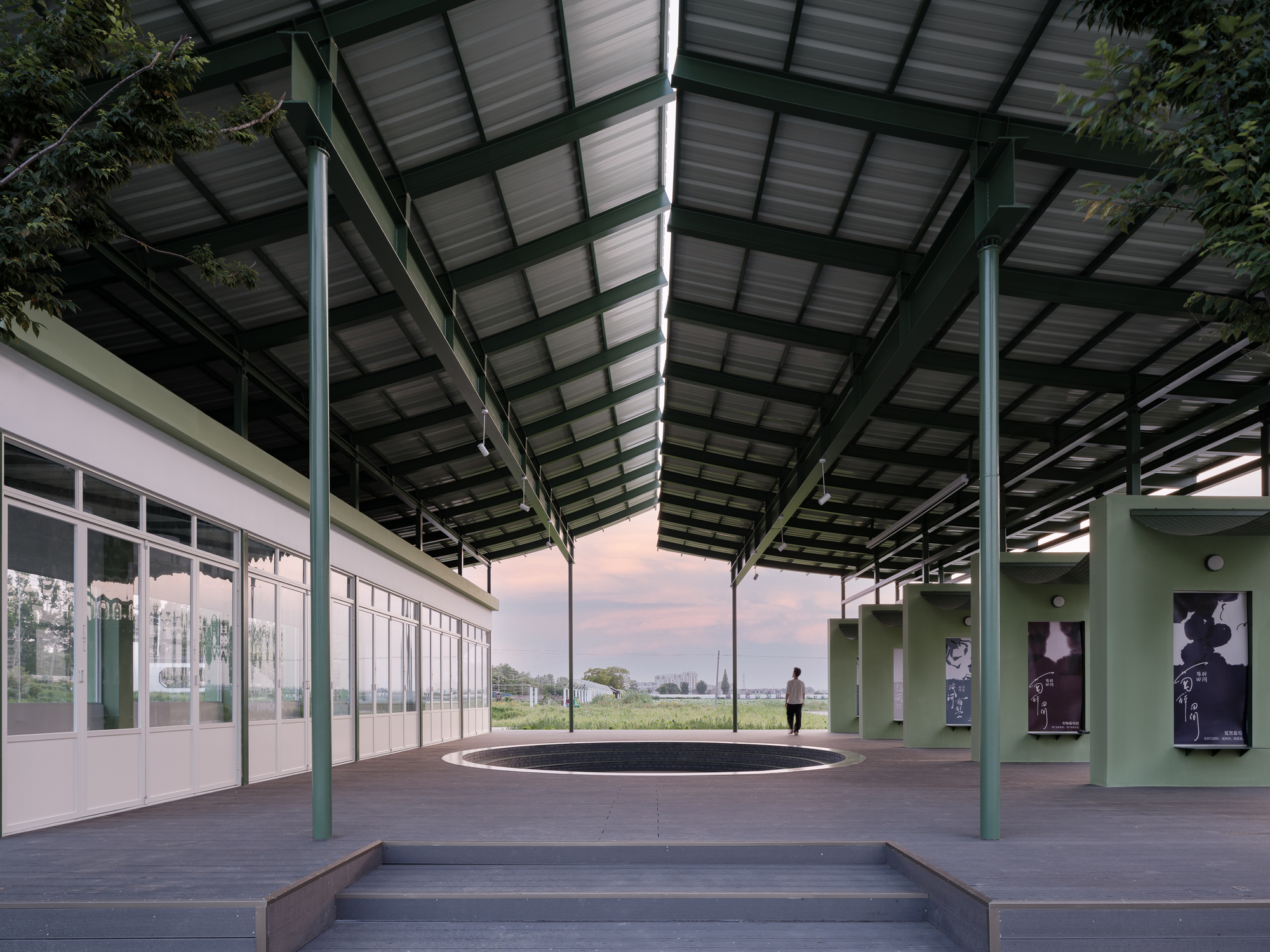 Covered outdoor platform with exposed steel structure, corrugated metal roof, and glazed classroom volume at dusk