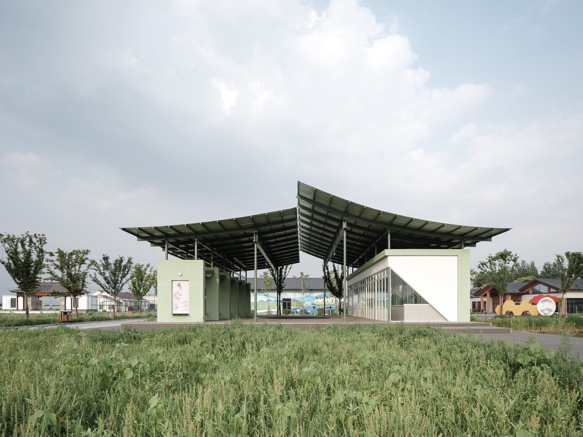 Pavilion with cantilevering metal roof sheltering green-painted volumes set in a grassy field under cloudy skies