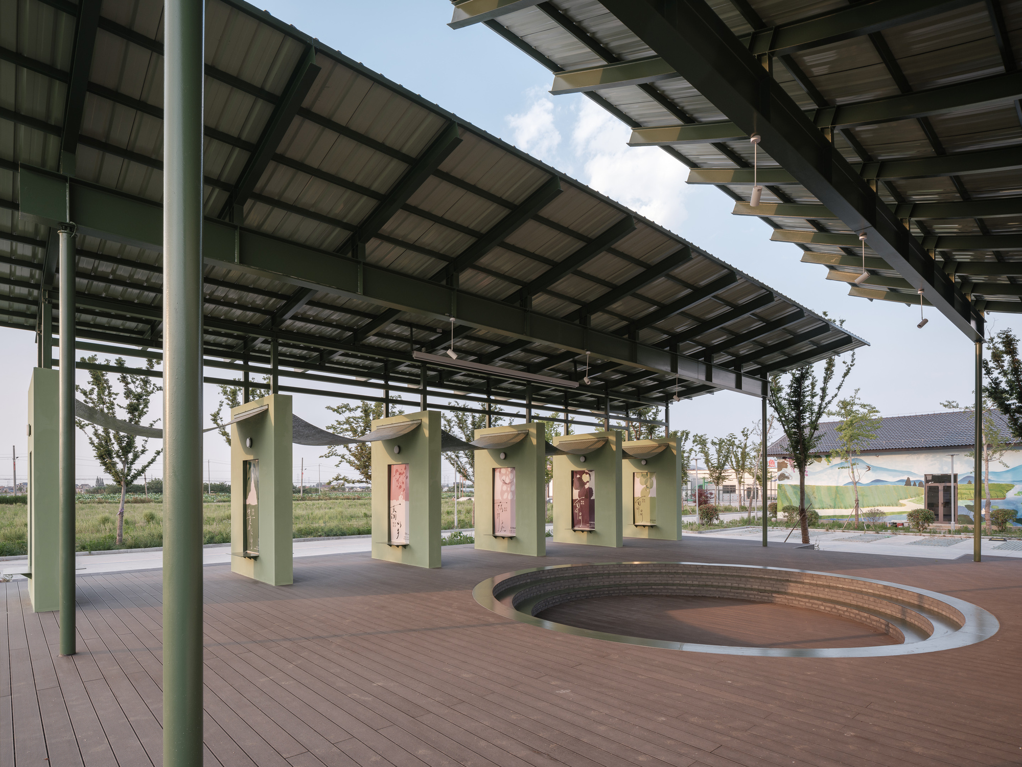 Open pavilion with solar panel roof and curved amphitheatre seating facing green exhibition kiosks