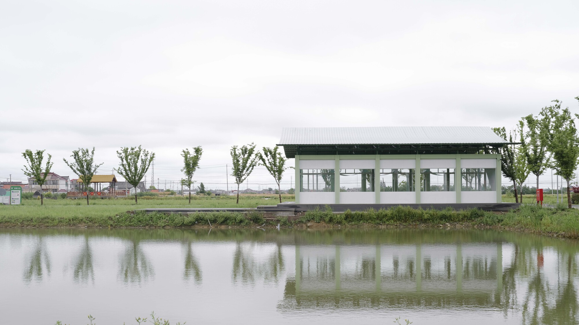 Glazed pavilion reflected in the still pond under overcast sky with row of young trees