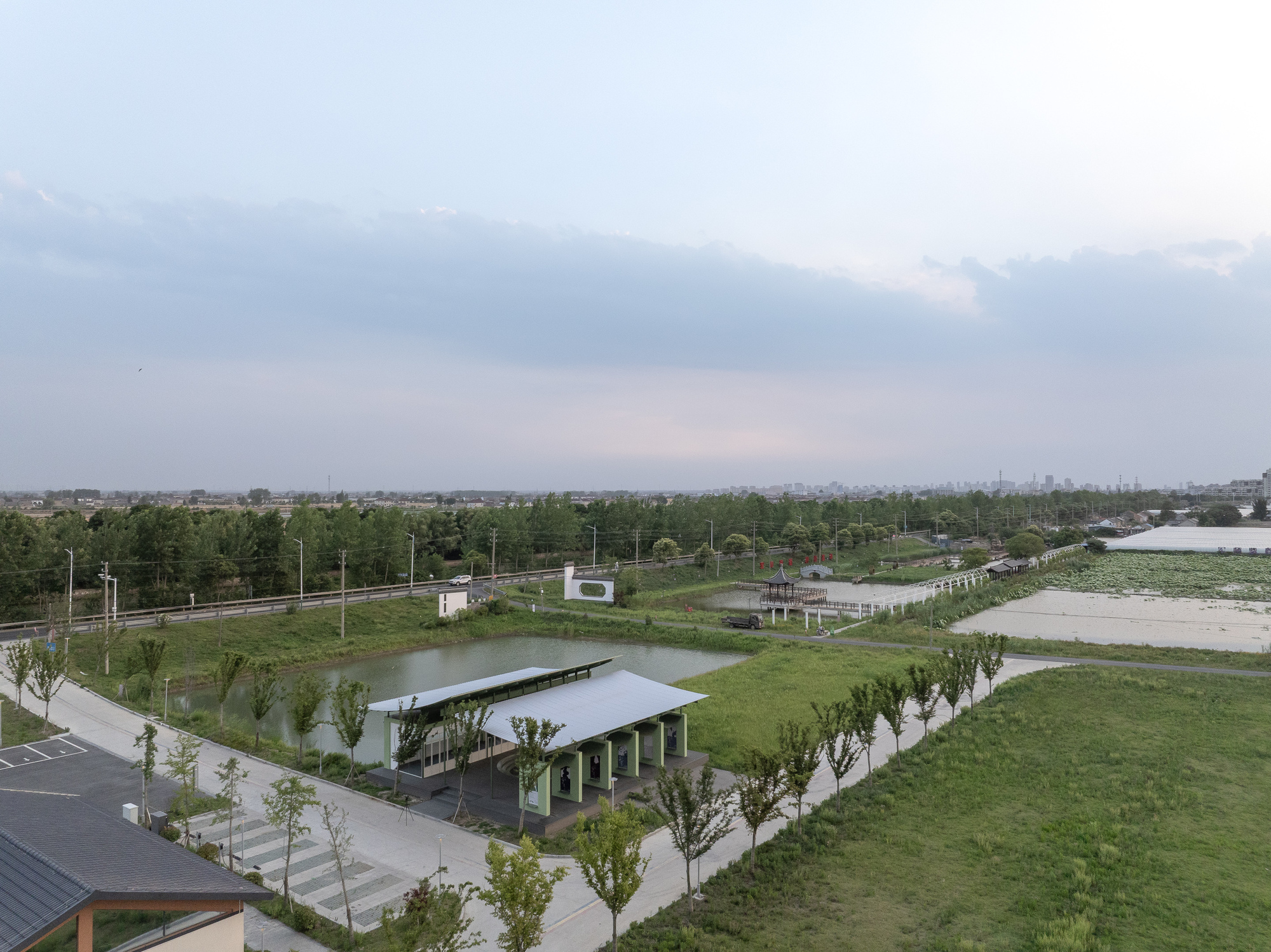 Elevated view of the pavilion with floating roof beside a pond and parking area under overcast skies