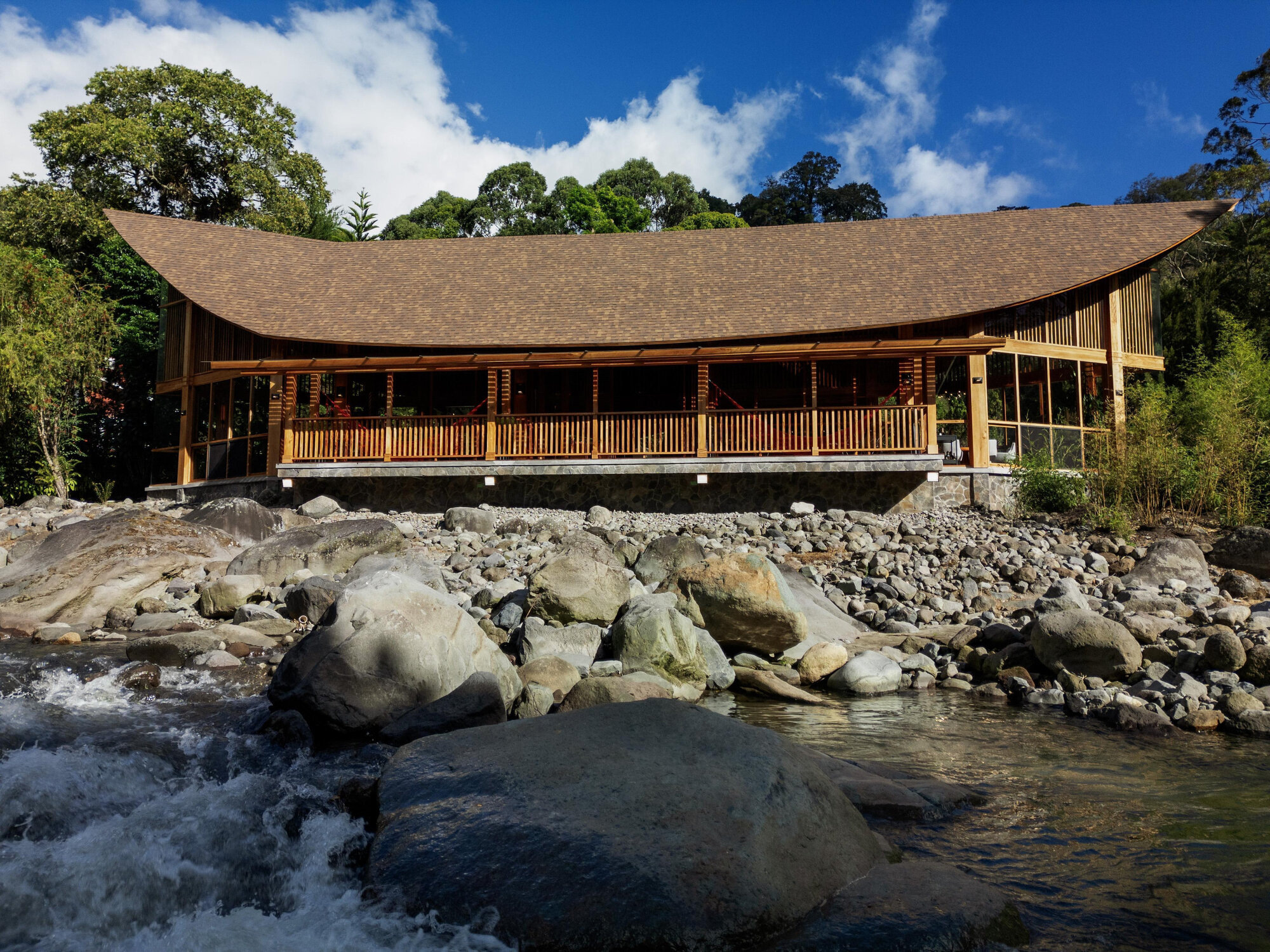 Timber pavilion with curved roof and slatted railings set above a rocky riverbed under blue sky