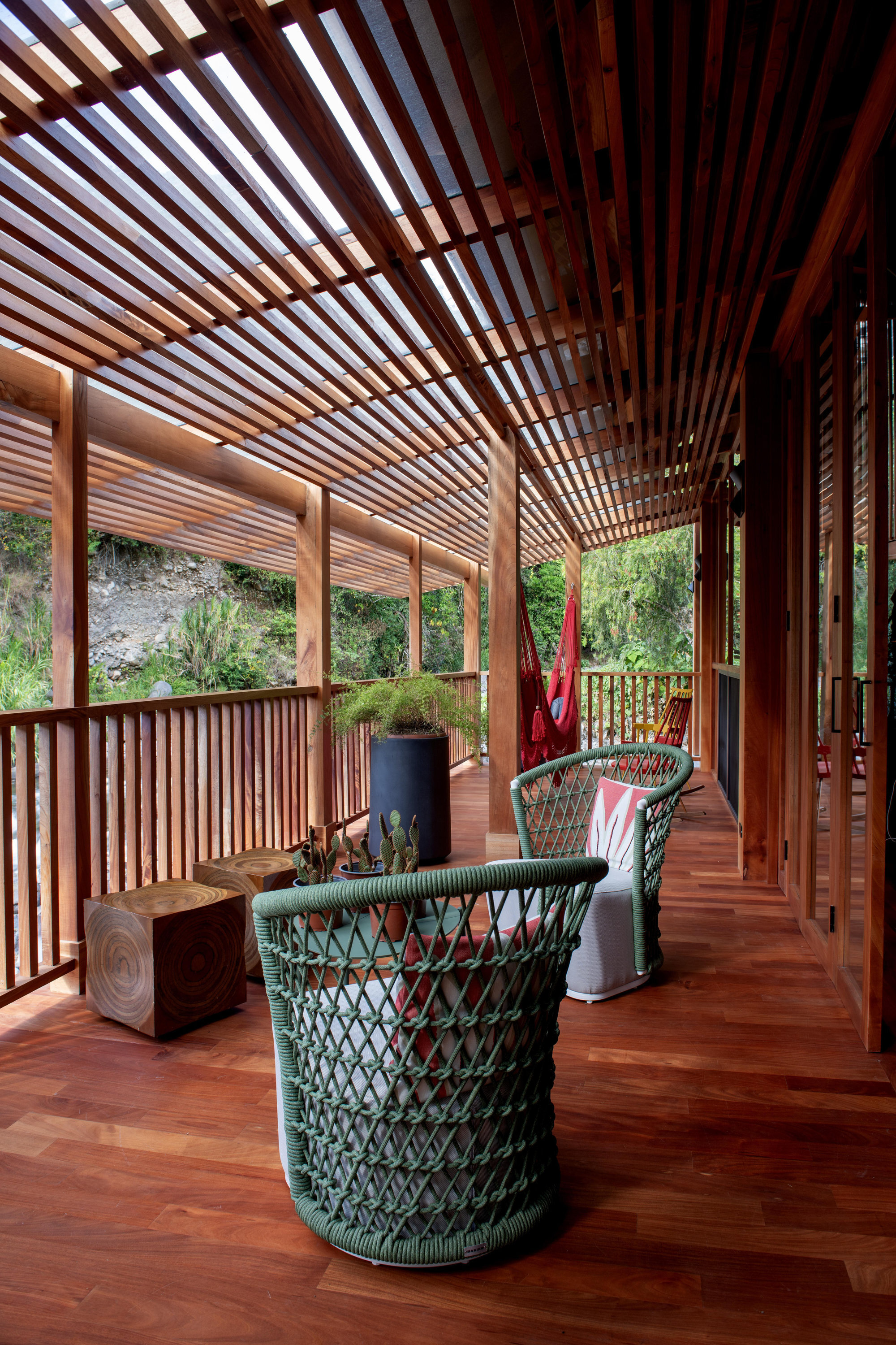 Covered terrace with slatted timber ceiling and railing overlooking hillside vegetation and woven chairs