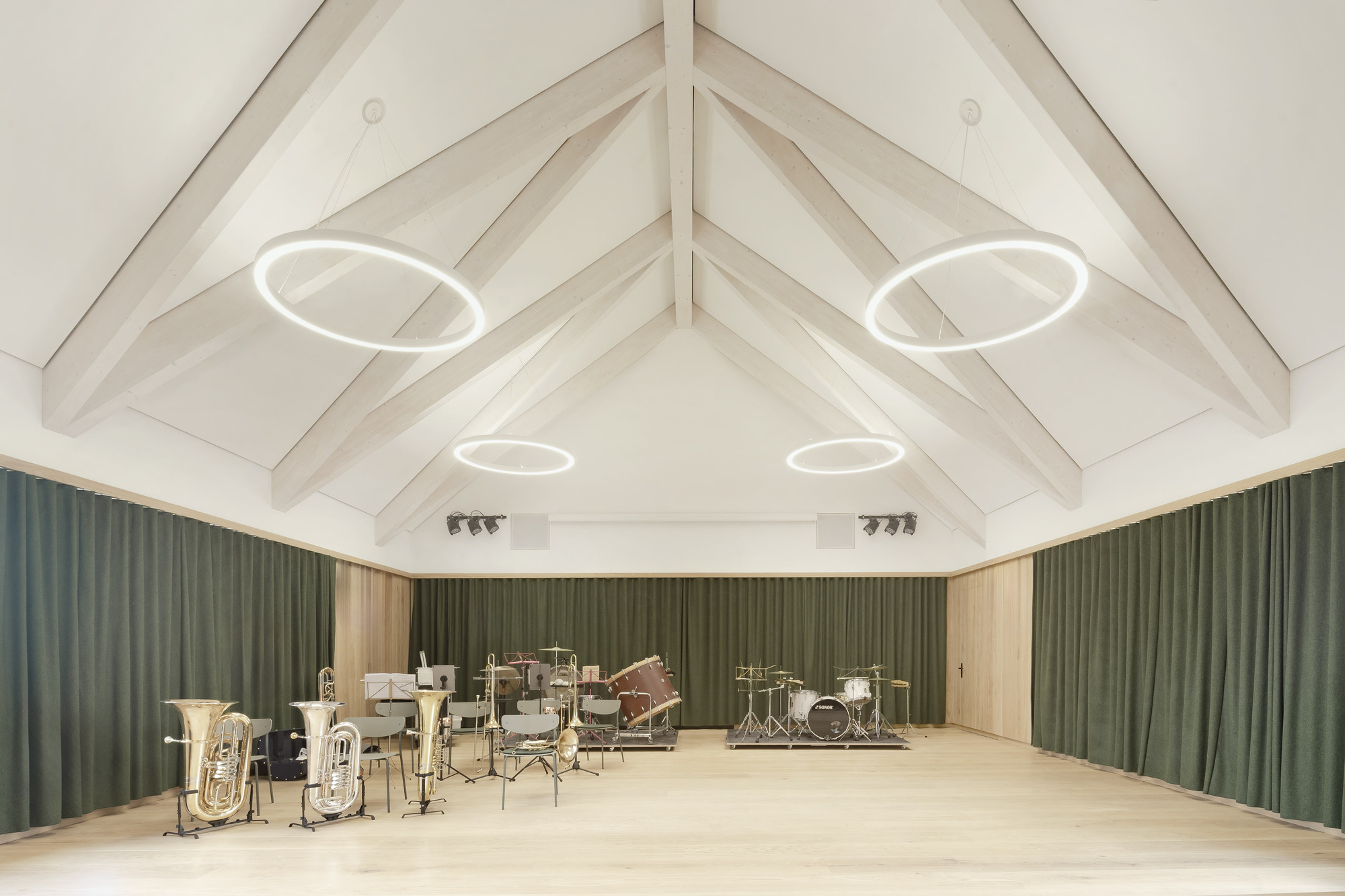 Rehearsal hall with exposed timber roof trusses, circular pendant lights, and green acoustic curtains surrounding brass instruments