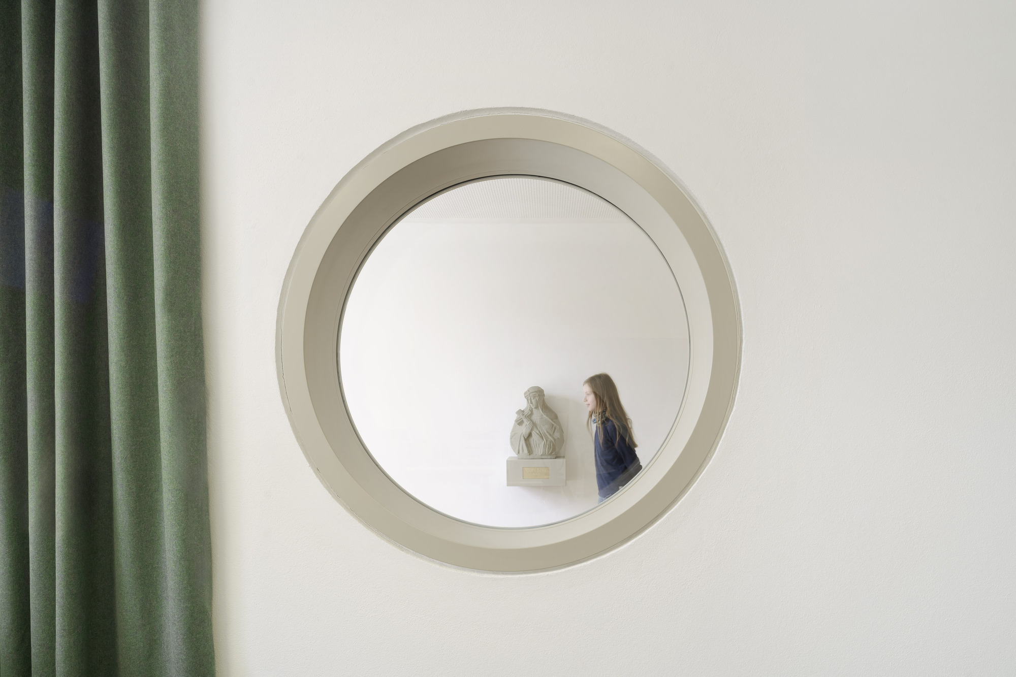 Circular window opening framing a visitor and a sculpture inside with green curtains visible at left