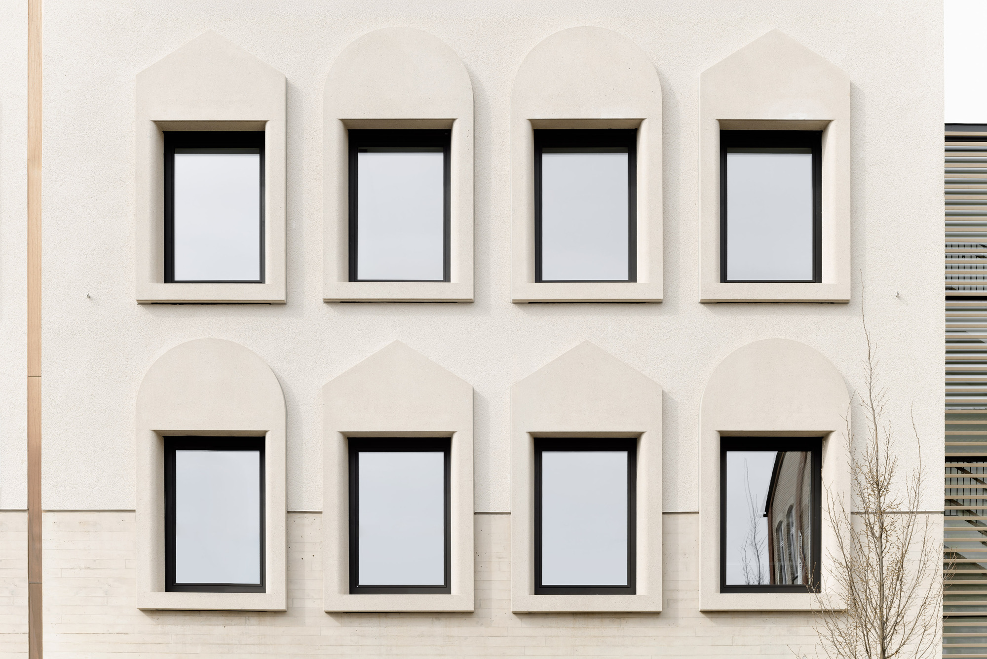 Facade detail of arched window openings with dark frames set in pale stucco beside a bare tree