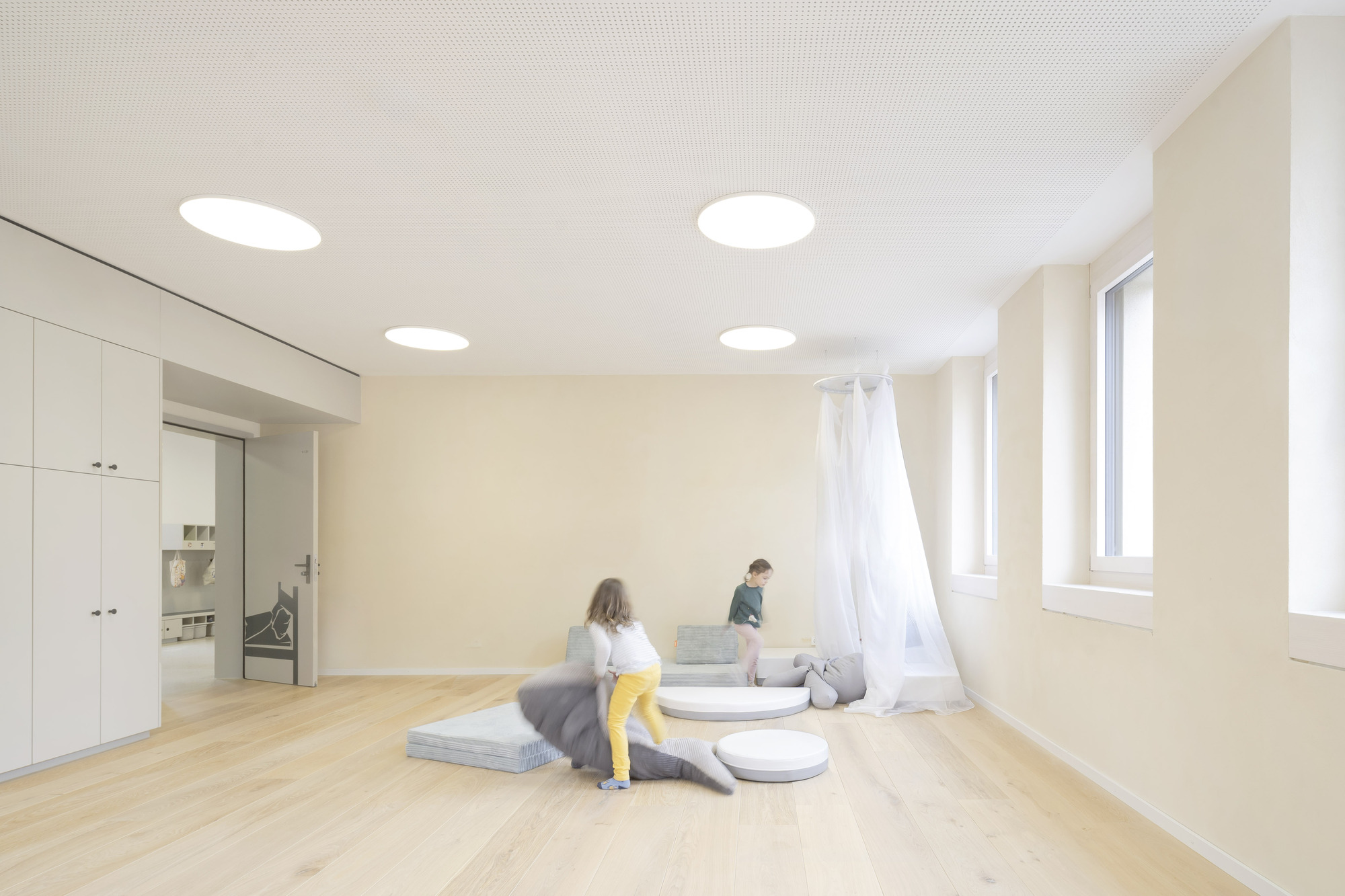Interior classroom with round ceiling lights, timber flooring, and children sitting on floor cushions near arched windows