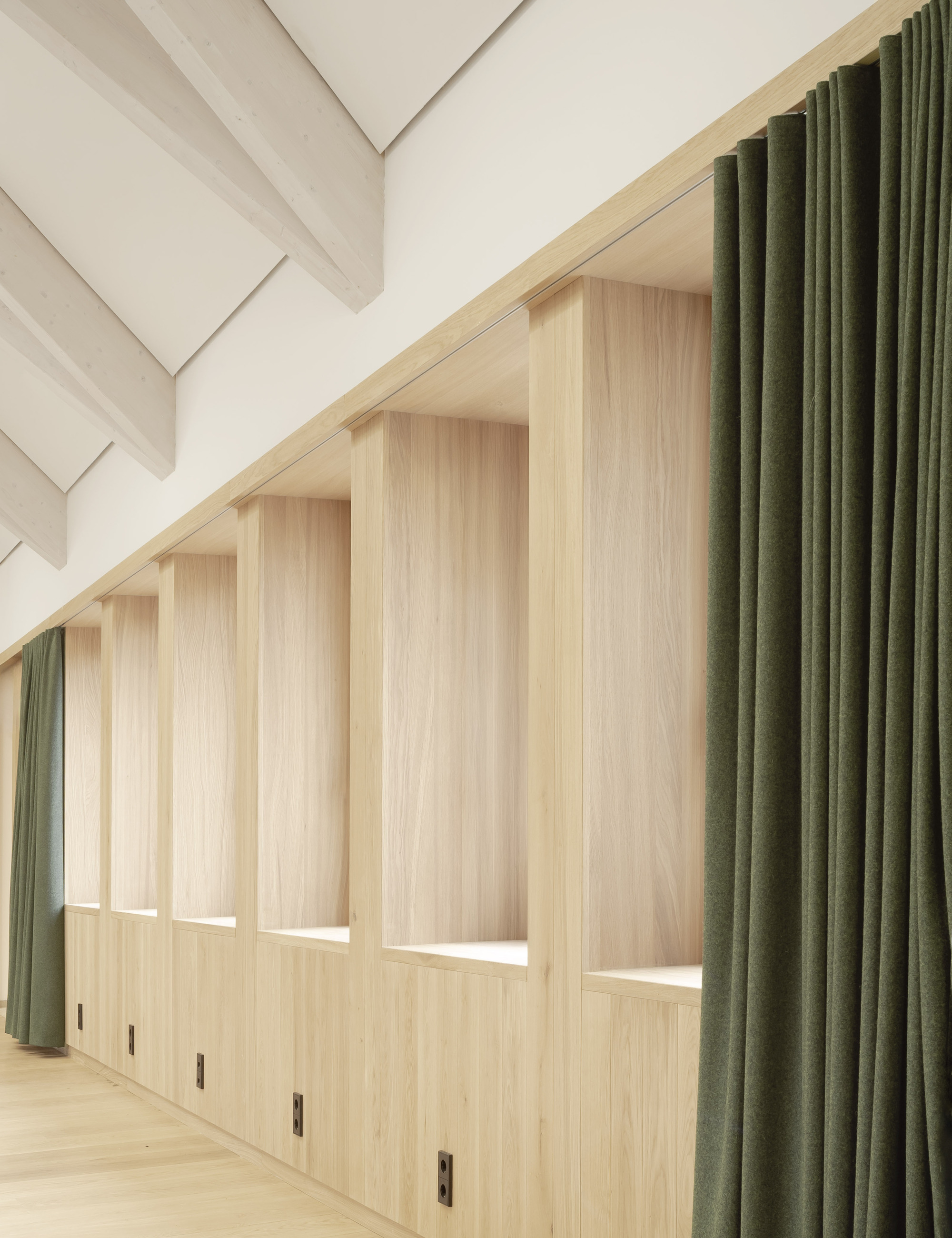 Light wood storage wall with integrated benches and olive green curtains beneath a sloped ceiling