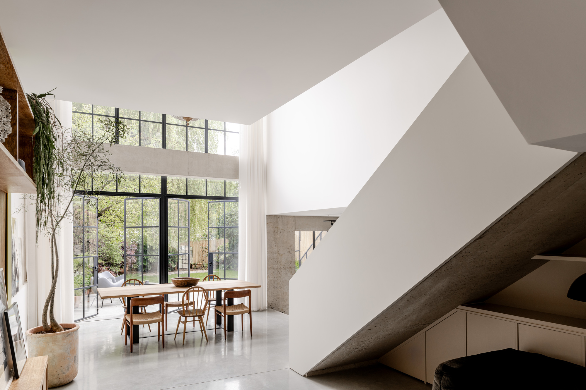 Double-height dining space with angled sculptural staircase and steel-framed glazing overlooking garden