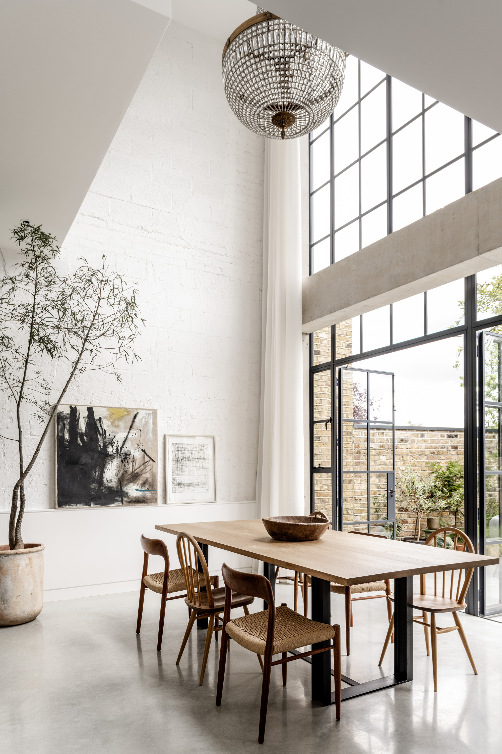 Double-height dining room with crystal chandelier and tall steel-framed windows framing courtyard trees