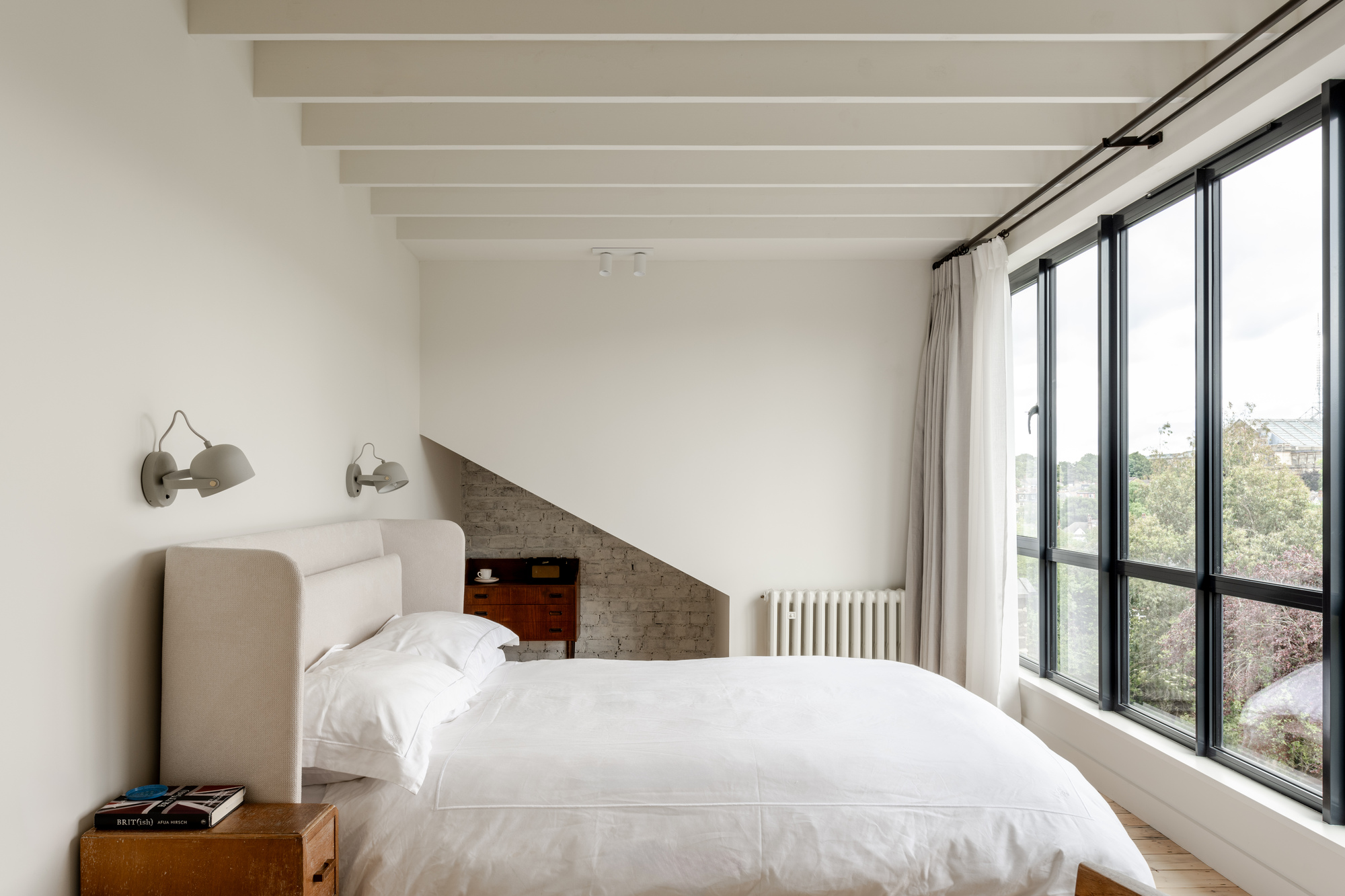 Bedroom with exposed ceiling beams and full-height steel-framed windows overlooking treetops