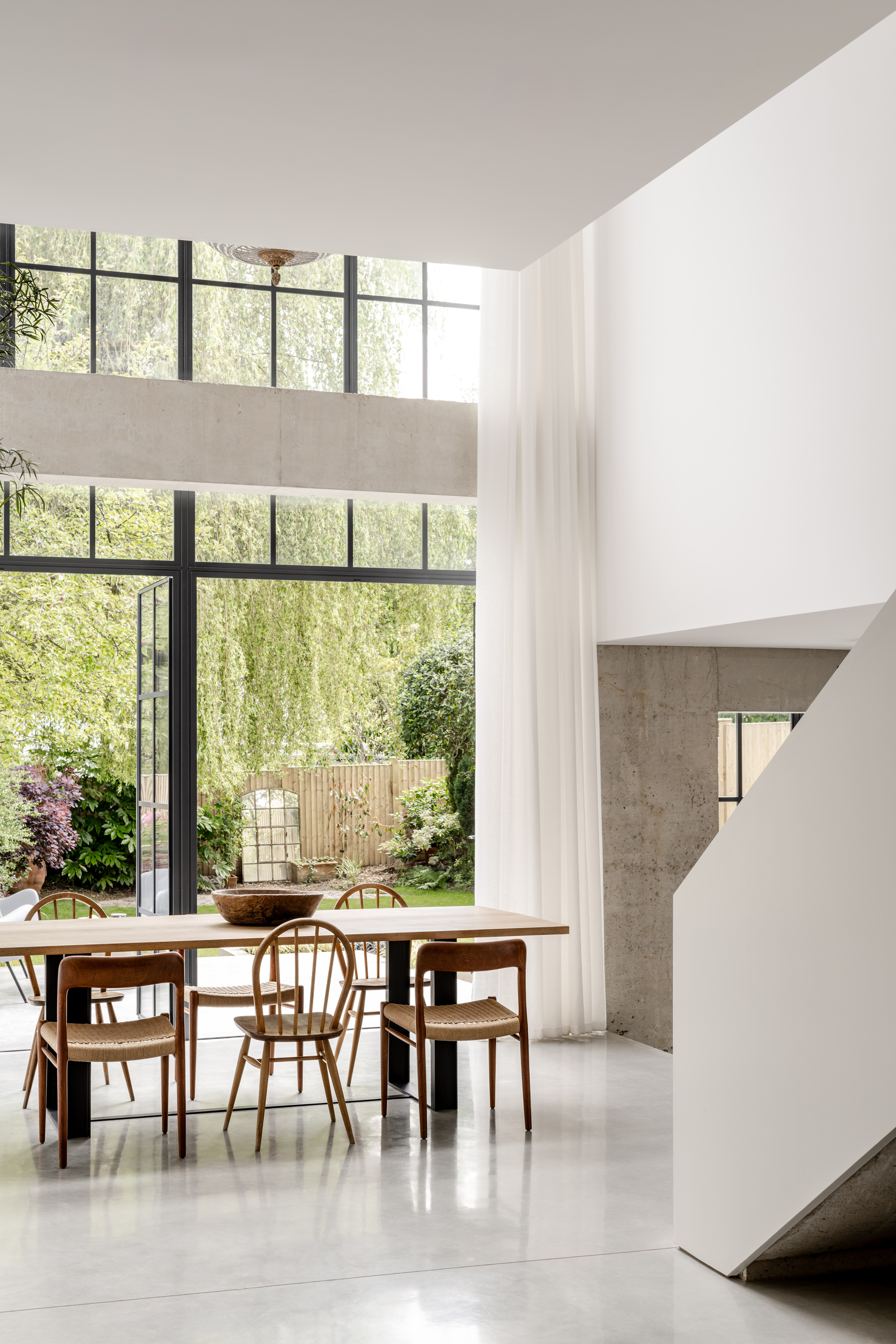 Dining area beneath double-height steel-framed windows overlooking a planted garden with willow trees