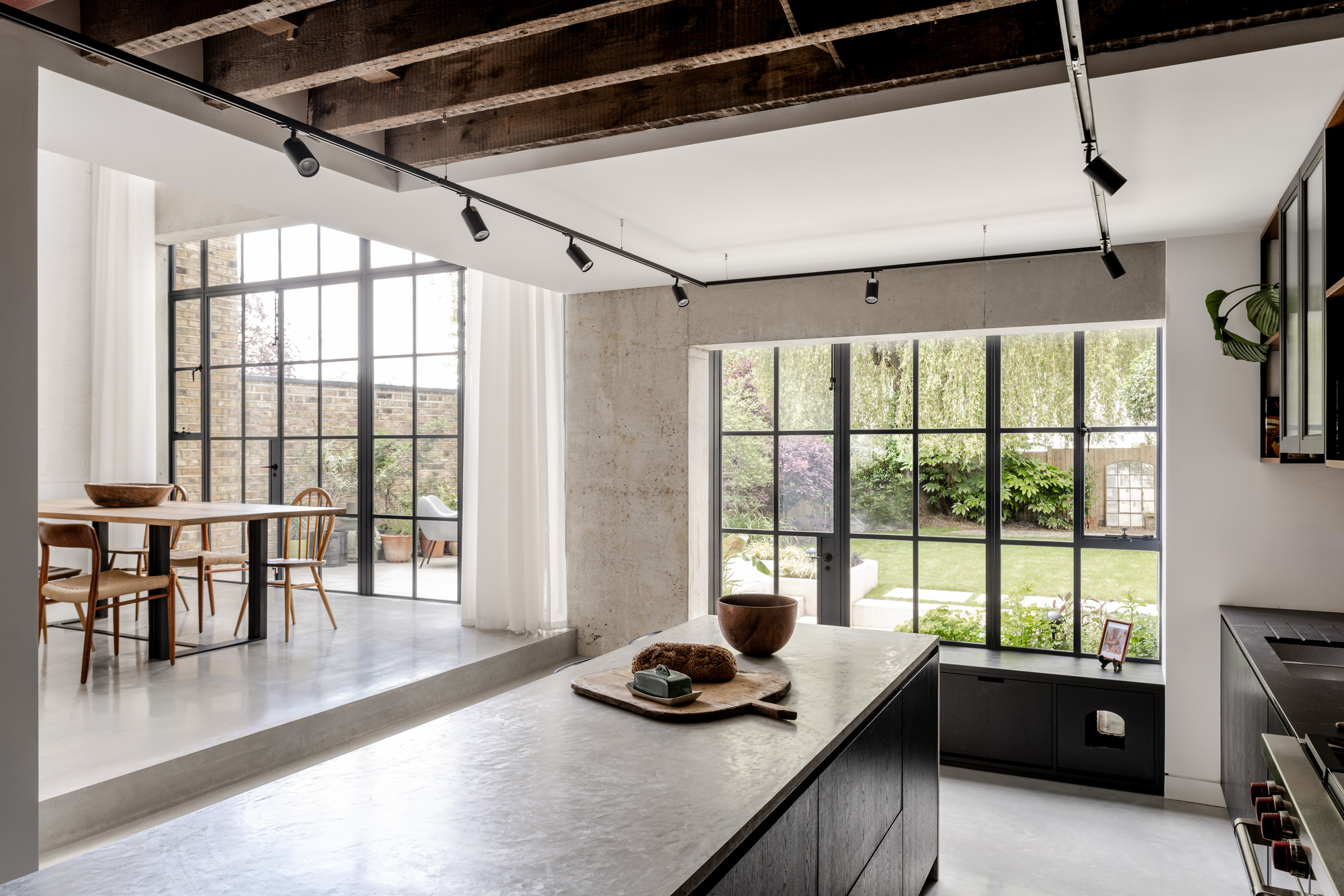 Kitchen with concrete island and exposed ducting beneath the ceiling looking toward steel-framed windows