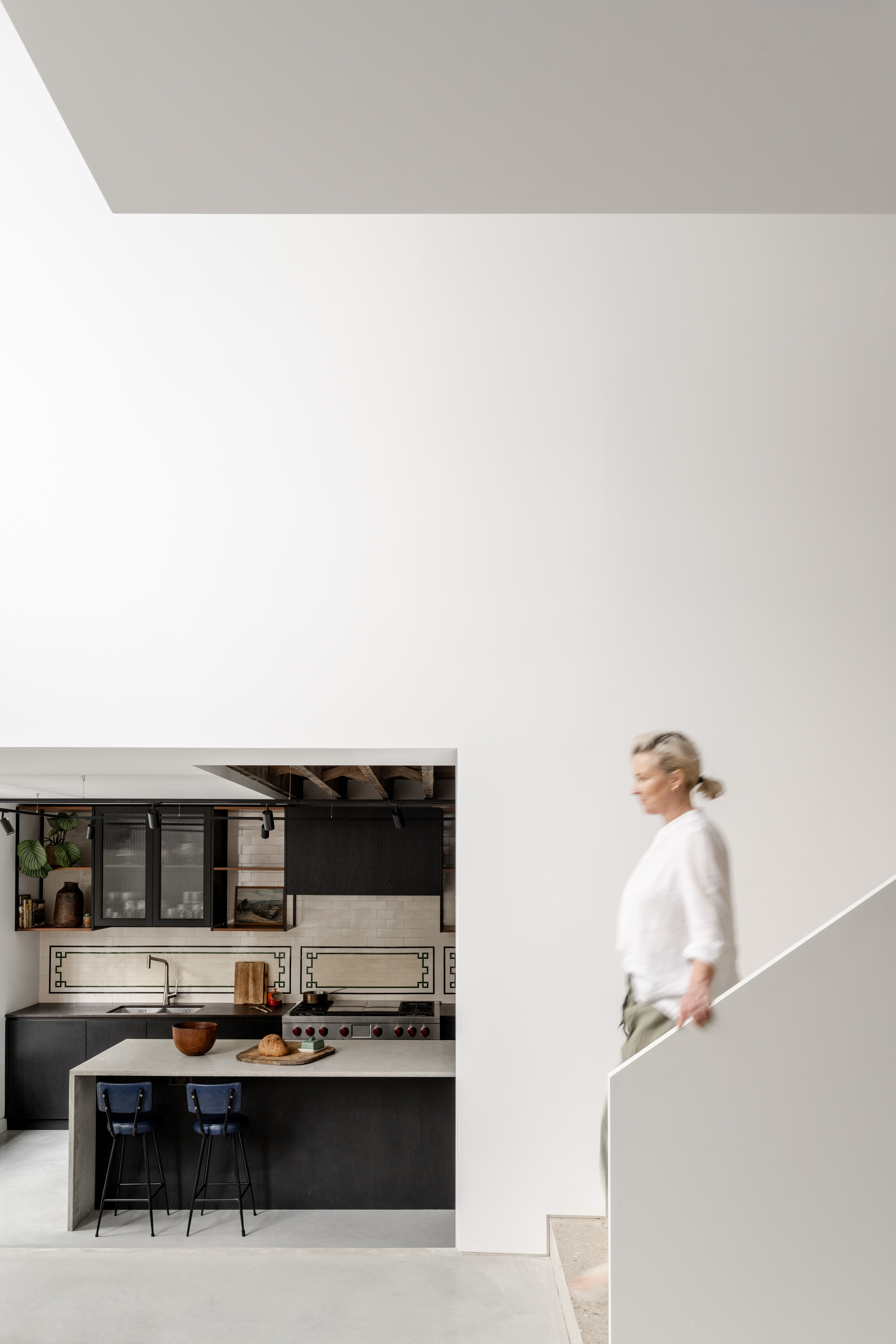 Person ascending a sculptural white staircase beside the black kitchen with exposed timber ceiling