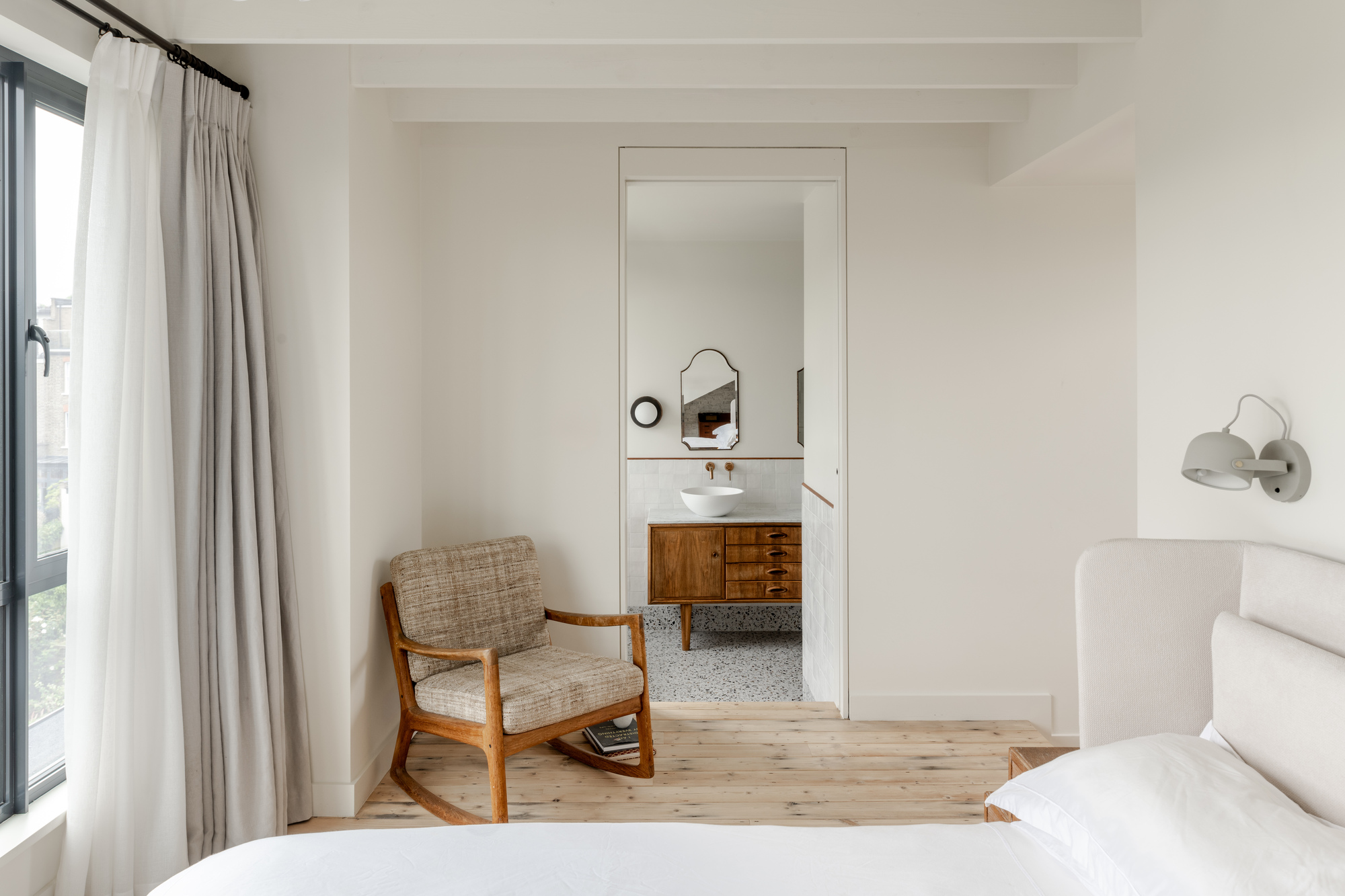 Bedroom with timber rocking chair looking through doorway to ensuite bathroom with terrazzo flooring