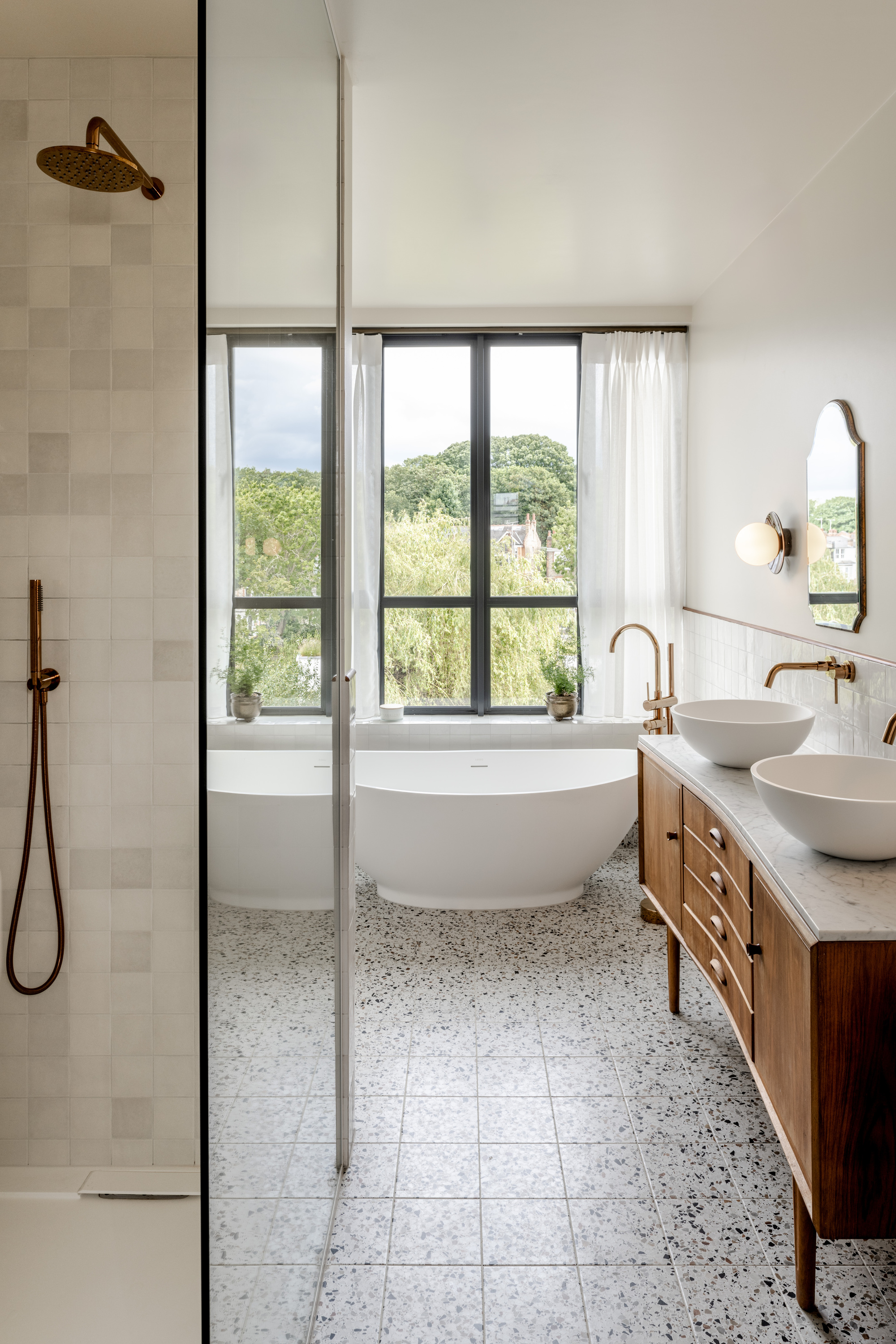 Bathroom with freestanding tub beneath steel-framed window overlooking trees and terrazzo floor throughout