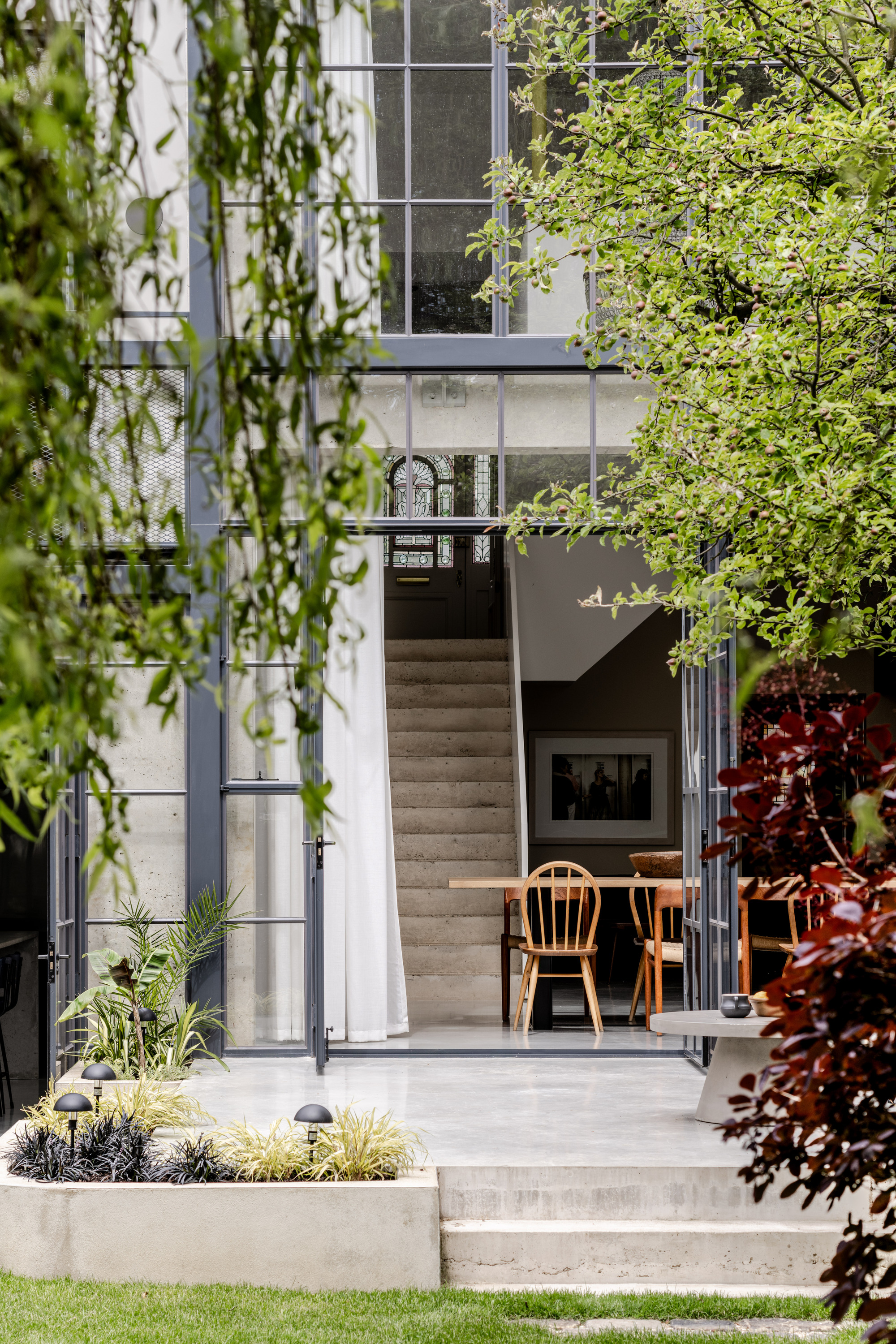 View through garden foliage to open glazed doors revealing a concrete staircase rising between interior and terrace