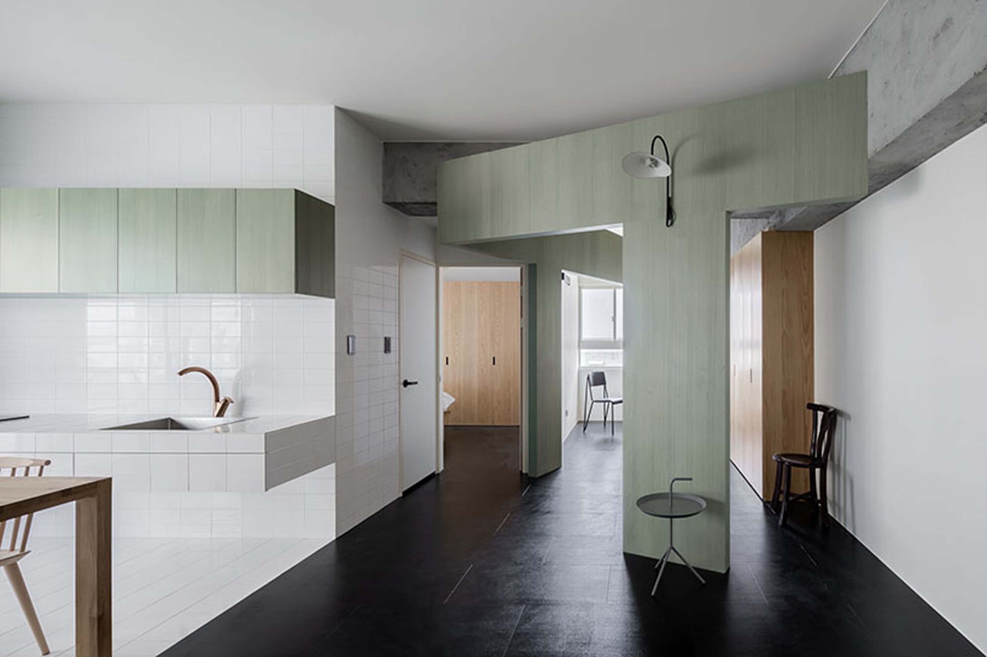 Open-plan interior with white kitchen island, pale green cabinetry, and black flooring under exposed concrete beams