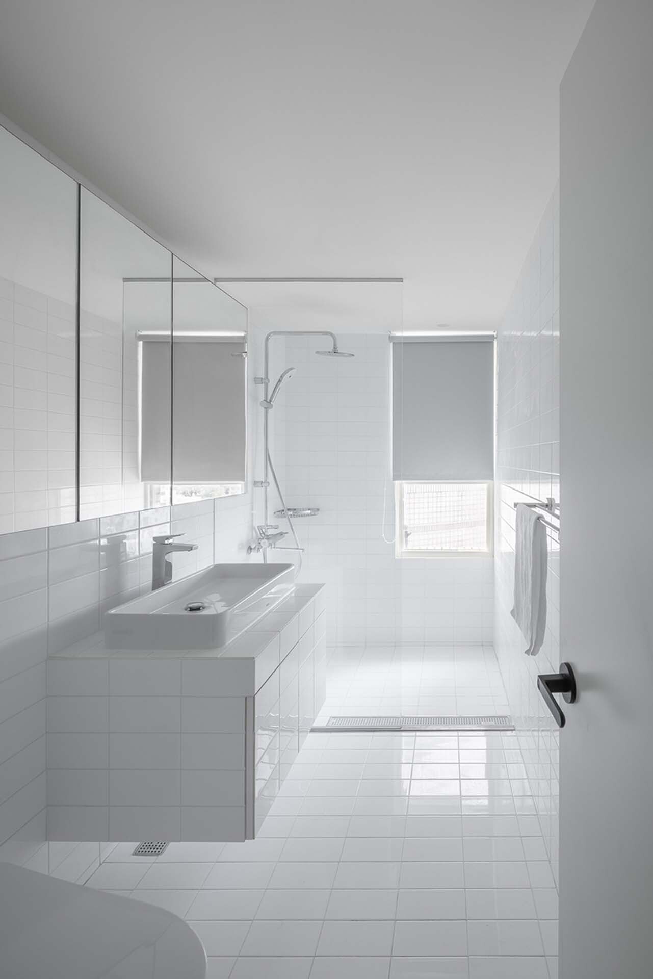 White-tiled bathroom with floating vanity, glass shower partition and windows with translucent blinds